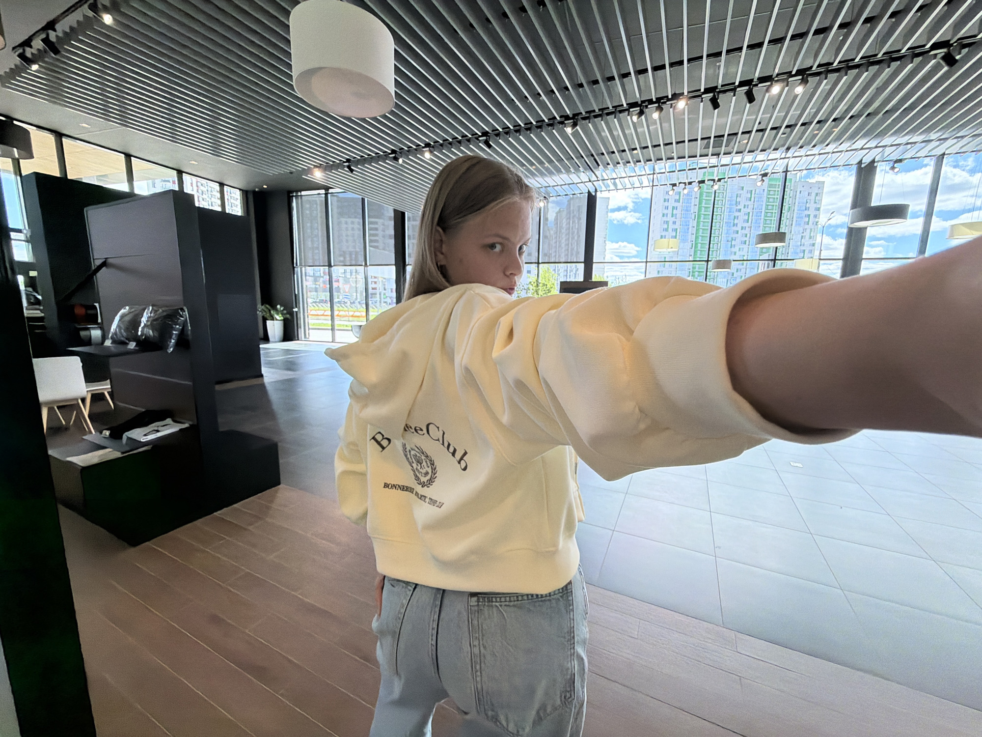A young, light-skinned person with blonde hair stands in a modern, well-lit interior space. The person is wearing a cream-colored hoodie with the words "Bree Club" and "BONNEIRET.EST.1997" printed on the back, paired with light blue jeans. They are facing away from the camera, with their head turned to the left, appearing to look back. One arm is outstretched toward the camera, creating a "selfie" perspective.
The background features a wall of windows overlooking a cityscape with tall buildings under a sky with scattered clouds. The interior space has a unique ceiling structure with parallel lines or beams running across it. There is also a modern lighting fixture hanging from the ceiling. In the foreground to the left, there is a dark-colored structure resembling a display or partition, along with a white chair. The floor appears to be a mix of light wood-grain planks and a white tiled surface.
Without precise location data, it is challenging to verify if the scene is located in Izhevsk, Russia. However, the architecture and general urban environment suggest it could be a contemporary office, showroom, or retail space in a modern city.