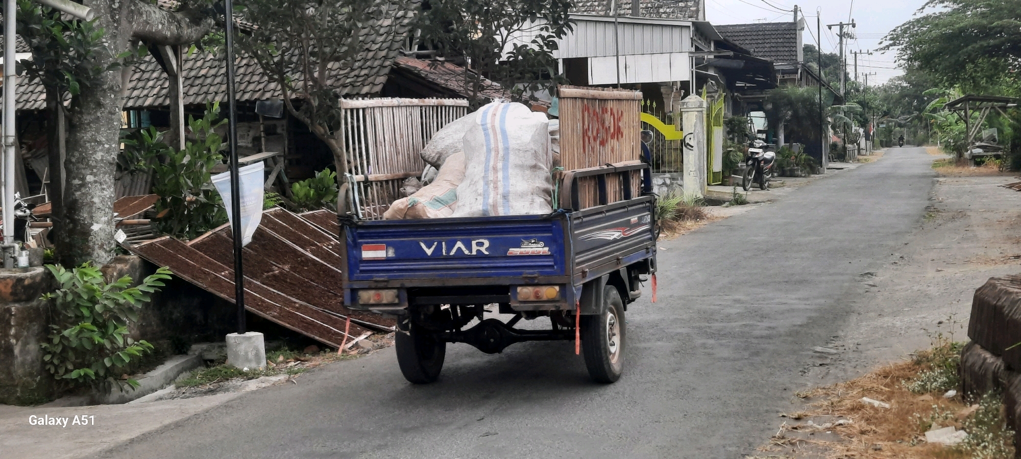 A blue flatbed truck with a white sack and a wooden crate filled with goods is parked on a paved road in Klemunan, Indonesia. The truck has the brand "VIAR" on its side. To the left of the truck, there is a house with a tiled roof and a wooden fence. In front of the house, there are some plants and a pile of what appears to be dried tobacco leaves. To the right of the truck, there is a residential area with more houses, a motorcycle parked in front of one house, and trees. The road continues into the distance, with more houses and vegetation visible on both sides. The sky is overcast. The image is a photograph taken from a slightly elevated angle, capturing the scene from the rear of the truck.