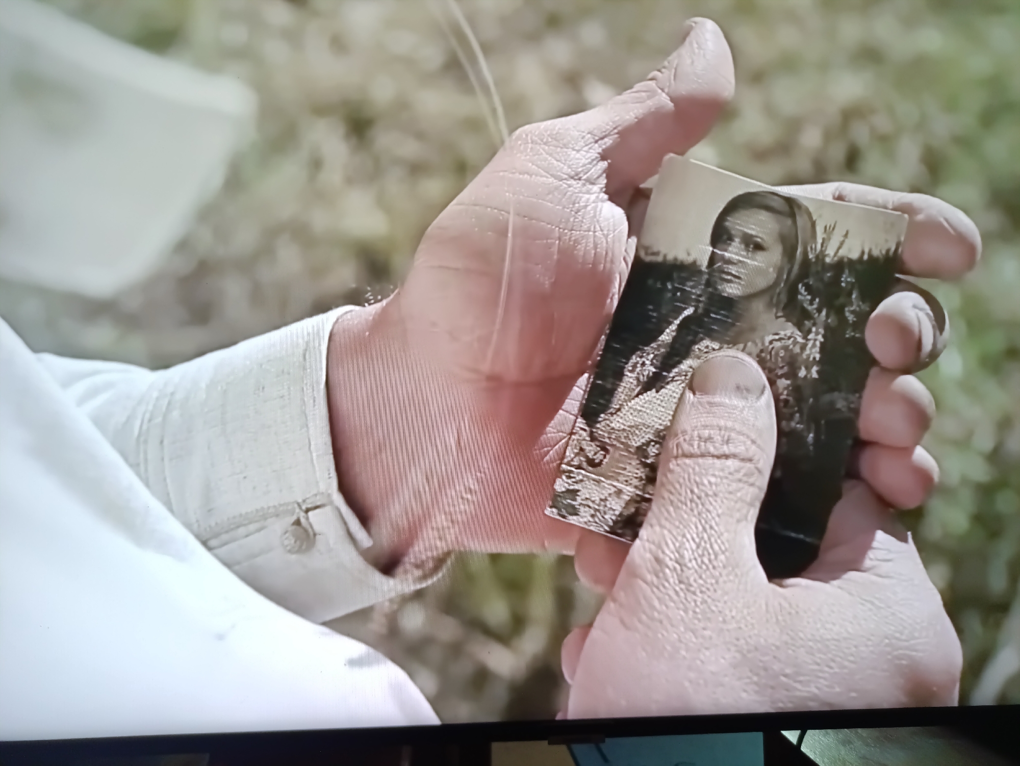 The image shows a close-up of a person's hands holding a small, vintage black and white photograph. The photograph depicts a young woman with her hair pulled back, looking directly at the camera. She is wearing a patterned dress, and the background of the photograph appears to be outdoors, possibly with foliage or tall grass.
The hands holding the photograph are weathered, suggesting an older individual. The person is wearing a light-colored, possibly linen, shirt with buttons visible on the cuff. The background of the scene is out of focus, showing a natural environment with muted green and brown tones, suggesting an outdoor setting during daylight hours. The lighting appears to be natural and somewhat bright, indicating it could be daytime.
The activity depicted is the examination of a photograph, which often evokes feelings of nostalgia, memory, or sentimentality. The photograph itself is worn, with some damage or creasing around the edges, further emphasizing its age and history.
No visible text is present in the image. The overall impression is one of personal reflection and remembrance, centered around a cherished photograph. The location context of Kamianets-Podilskyi, Ukraine, adds a layer of potential historical or personal significance to the image, though this cannot be directly inferred from the visual alone.