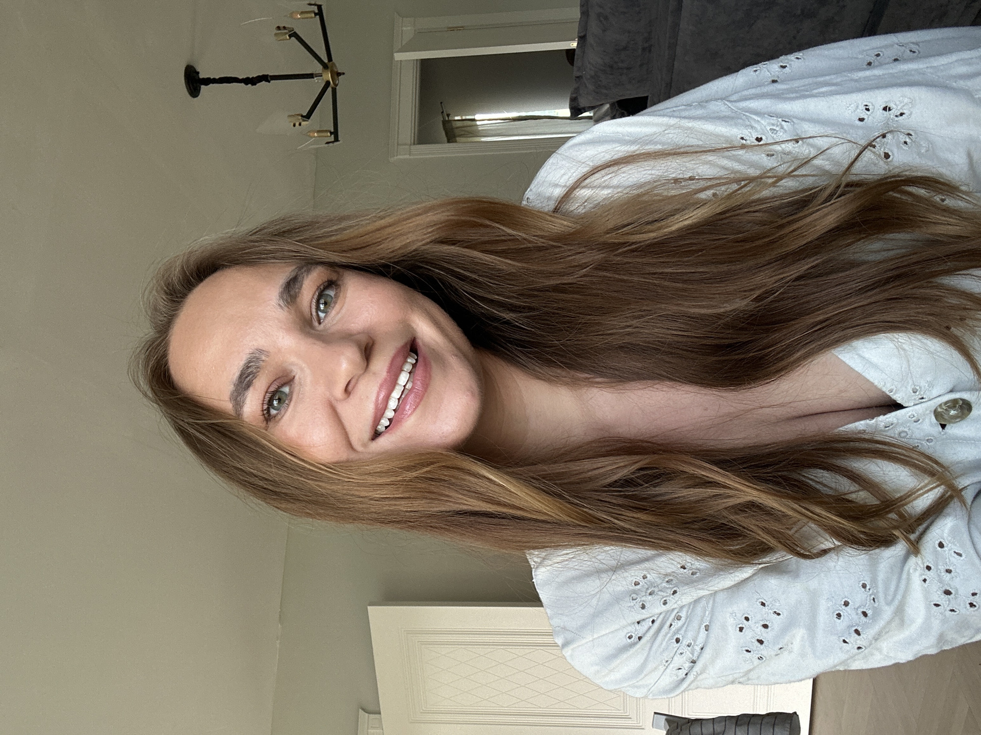 This is a close-up, vertically oriented selfie of a young woman with long, light brown hair and green eyes. She is smiling, showing her teeth, and her hair is partially obscuring her face. She is wearing a white, eyelet-detailed blouse with visible buttons. The background is indistinct, but appears to be indoors, possibly a bedroom, with a chandelier visible on the ceiling and a doorway or closet in the lower right. The lighting suggests daytime. The overall mood is happy and relaxed. There is no visible text in the image. The context provided is "Kharnavy, Russia".