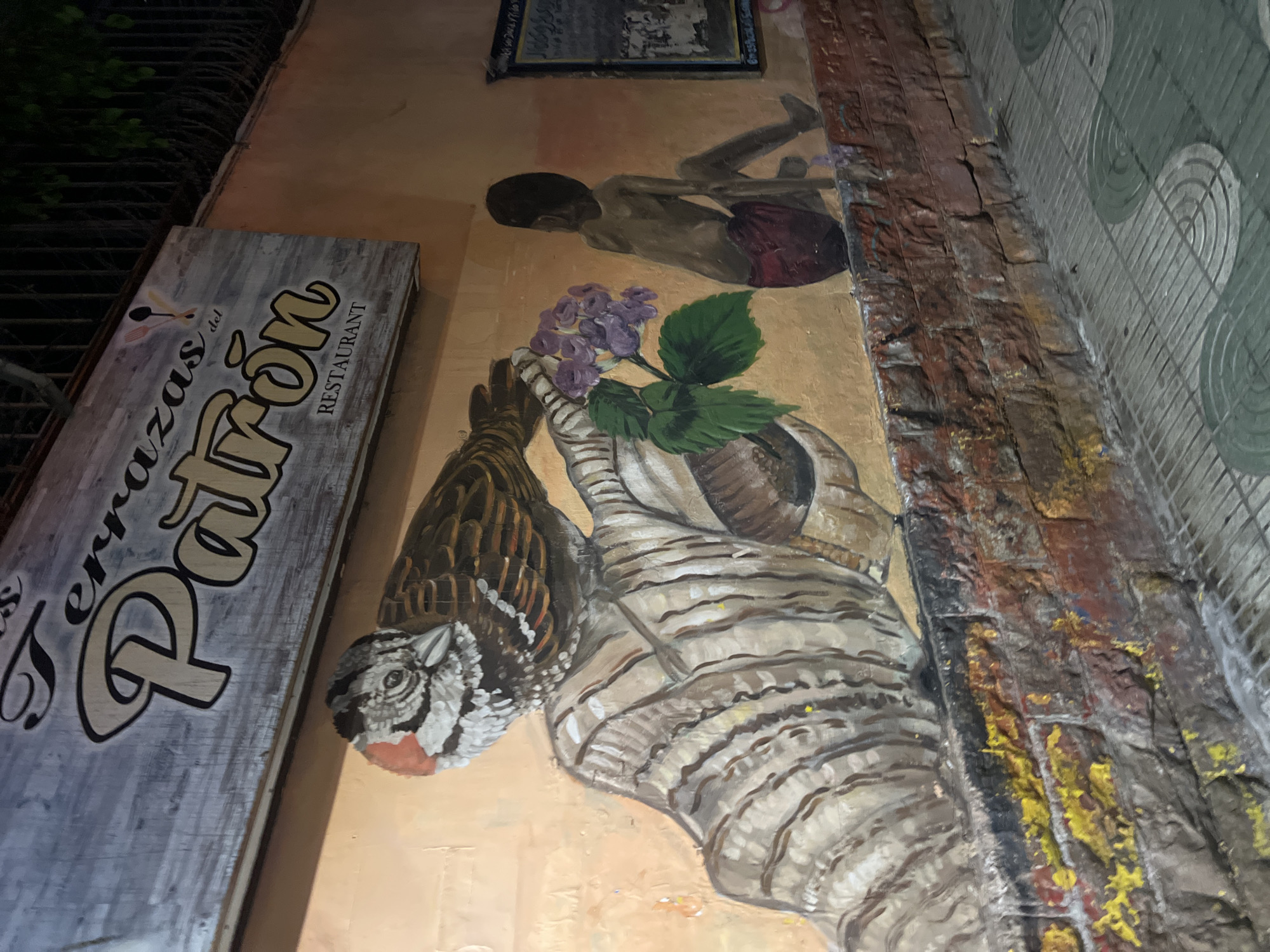 A street-level, slightly tilted medium shot presents a mural on an exterior wall in Arica, Chile. The mural is executed in a naive style and features several elements. At the top, a painting of a dark-skinned figure wearing red shorts is visible. Below that, there's an image of a striped animal, possibly a feline, holding purple flowers. Beside the mural, on the left, a wooden sign advertises "Terrazas del Patron" Restaurant. The adjacent structure on the right is a brick wall merging into a tiled surface, likely a sidewalk. The overall environment suggests a narrow street or alleyway.