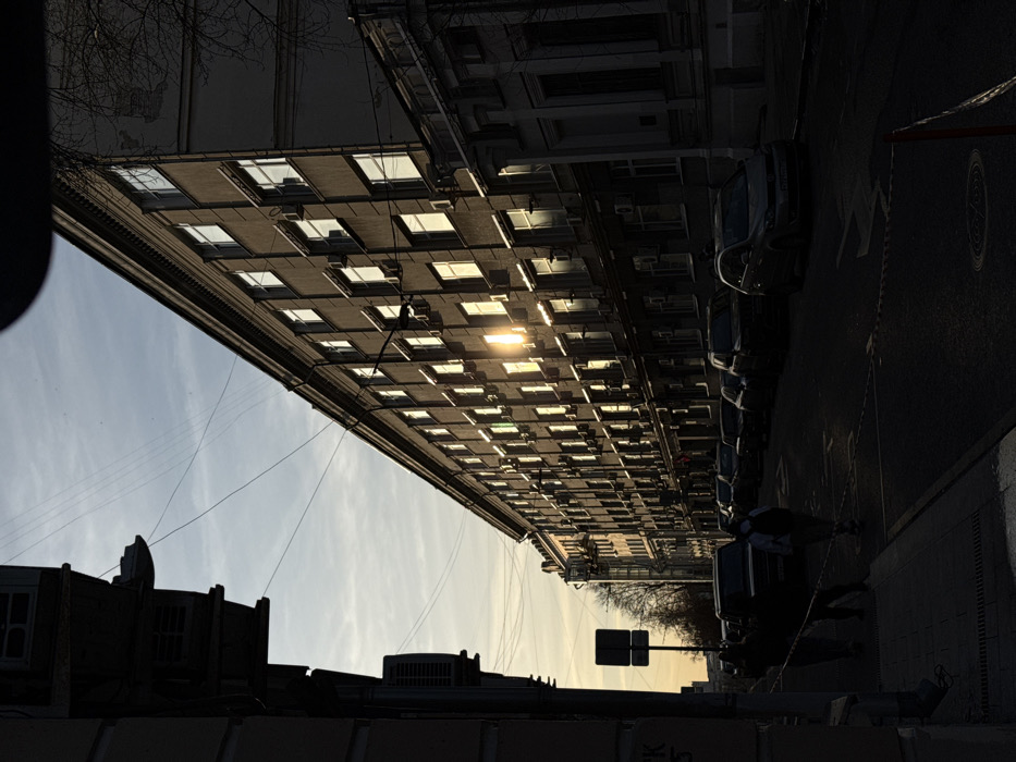 The image depicts an urban street scene in Moscow, Russia, specifically along Bolshoy Savvinsky Lane, with a low sun angle suggesting late afternoon or early morning.
On the right, a long, multi-story building (at least six visible floors) with a dark grey or beige facade extends into the distance. Numerous rectangular windows, many equipped with external air conditioning units, intensely reflect bright, low-angle sunlight. A prominent cornice defines the building's roofline.
The street, appearing dark due to heavy shadow, is paved with asphalt. Several vehicles, including sedans, SUVs, and a lighter-colored hatchback, are parked along the right side of the road. Two sections of white and red striped barriers or tape are present on the asphalt in the foreground. A circular manhole cover is visible on the street surface.
On the left, a heavily shadowed, narrower building is partially visible. A sign with the Cyrillic letters "ПК" and the numeral "5" is faintly discernible on its wall. Overhead utility wires crisscross the light blue sky, which contains sparse clouds.
Three individuals are visible in the mid-ground, heavily silhouetted by the shadow. Two figures are walking together on the sidewalk to the far left, while a third individual, possibly carrying a backpack, walks on the street towards the viewer, slightly right of the pair. A dark, rectangular street sign on a pole stands on the left side of the street, past the figures. Bare tree branches are visible against the sky in the upper right corner.