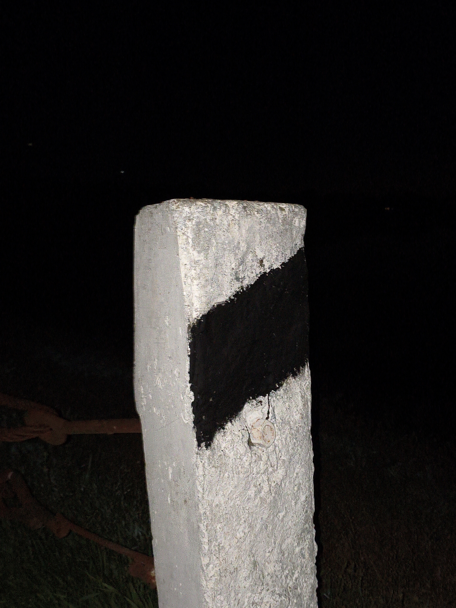 The image features a weathered, rectangular concrete or stone post standing vertically, centered in the frame against a predominantly dark background. The post is painted white and has a distinct diagonal black stripe extending from its upper left to its lower right. Its surface is rough and textured.
Near the bottom right edge of the black stripe, a small, light-colored, round object, possibly a snail shell or a similar natural attachment, is visible on the post. A small spider can be seen on the upper left portion of the post, above the black stripe.
The setting appears to be outdoors at night, indicated by the almost entirely black background, which suggests a lack of ambient light. Faint, distant specks of light are barely discernible in the darkness. In the lower left quadrant of the image, partially obscured by the post, there are hints of rusty metal components, possibly part of a chain or fence, and some dark ground cover, likely grass or vegetation.
The post itself resembles a roadside marker or a boundary post often found along roads or property lines. There are no people visible, and no specific activity is taking place. No text is discernible anywhere in the image.