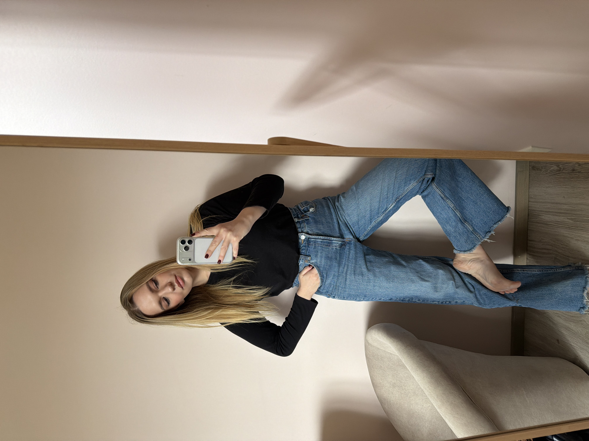 A young, fair-skinned woman with long, light brown to blonde hair takes a mirror selfie in an indoor setting. She is wearing a black long-sleeved shirt and blue jeans. She holds a smartphone with a white case in her right hand, the camera facing her. Her left hand is in her jeans pocket. She is barefoot. The background includes a light pink wall, a light beige chair, and a wooden-framed mirror. The floor appears to be wood-like. The location is likely an apartment or house, but its exact address or location within Dreiliņi, Latvia, cannot be determined from the image.
