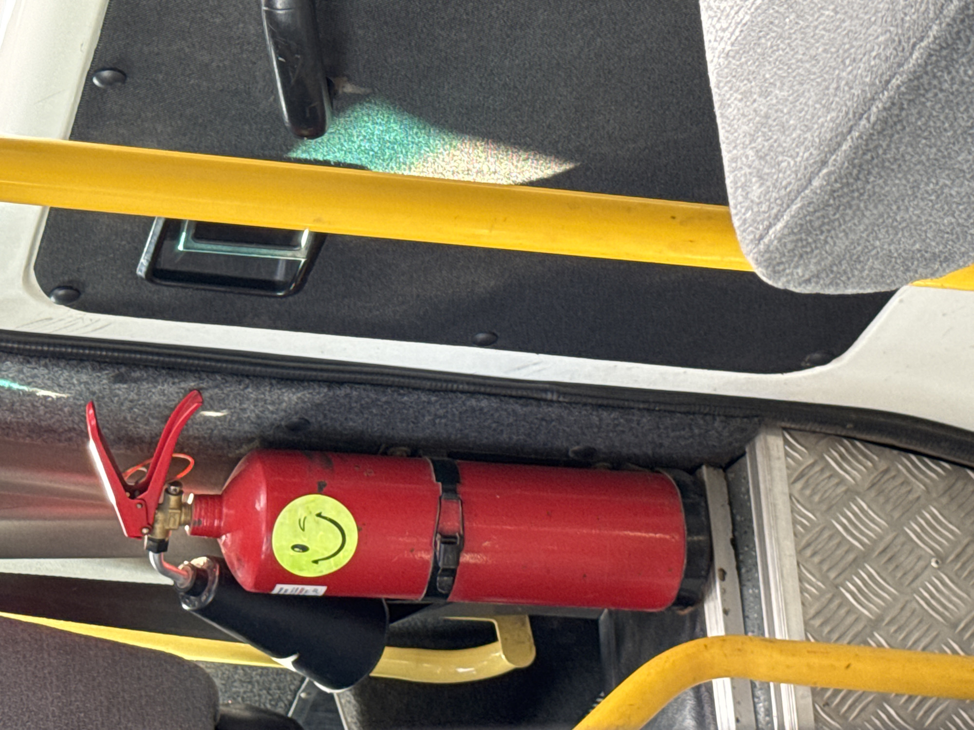 This media file is a close-up, eye-level shot of a red fire extinguisher mounted horizontally within what appears to be the interior of a vehicle, possibly a bus or van. The fire extinguisher is prominently displayed with a yellow smiley face sticker on its side. It has a red handle with a black lever and a metal nozzle.
The setting shows a textured dark grey surface above the fire extinguisher, with a bright yellow horizontal bar running across the upper portion of the frame, partially obscuring a reflective, iridescent patch of light on the dark surface. To the right of the fire extinguisher, a textured grey seatback is visible. Below the fire extinguisher, a yellow bar curves out of view. The flooring or lower surface of the vehicle shows a metallic, diamond-plate texture.
There is no visible activity or event taking place; the image is a static capture of the equipment. The lighting suggests an indoor environment, possibly during the daytime given the bright yellow bar and the light reflection. The location cues, such as the type of interior and the presence of a fire extinguisher, suggest a public transport or commercial vehicle. No text is clearly legible on the fire extinguisher or in the surrounding environment, though there might be small markings on the sticker. The overall impression is one of preparedness and safety equipment within a functional vehicle.