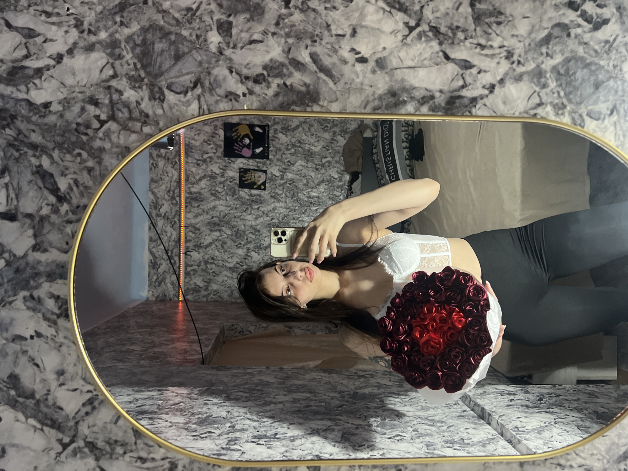 A young woman is taking a selfie in a mirror. She is wearing a white lace bra and black leggings. She is holding a bouquet of red roses. The background is a wall with a marble-like pattern. The reflection in the mirror shows more of the room, including posters on the wall and some furniture. The location is inside a building in Blizhnii Khutor, Moldova.