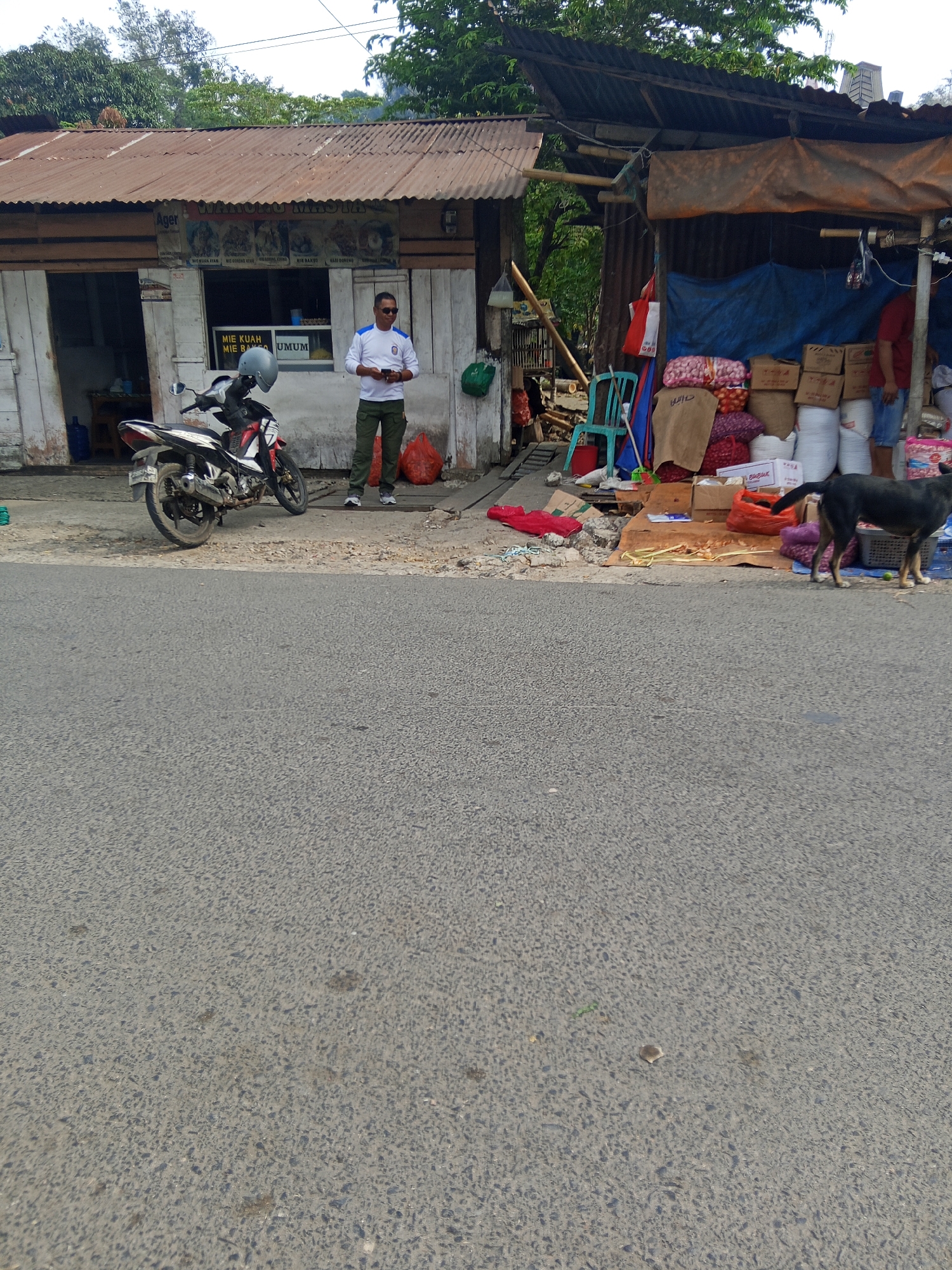 In the city of Makale, Indonesia, a man stands on a dirt path in front of a small, open-fronted building with a corrugated metal roof. The building appears to be a shop. A motorcycle is parked to the left of the man. The shop's facade has several windows and signs, including one with the text "MIE KUAH MIE BASO UMUM" and another with "Agar".
To the right of the shop, a makeshift stall is set up with tarps and bamboo poles. Various goods, including sacks of onions, bags of white material, and cardboard boxes, are displayed for sale. A dark-colored dog stands near the stall. In the foreground, a paved road runs horizontally across the image. The overall environment suggests a roadside market or small business in a semi-urban or rural setting.