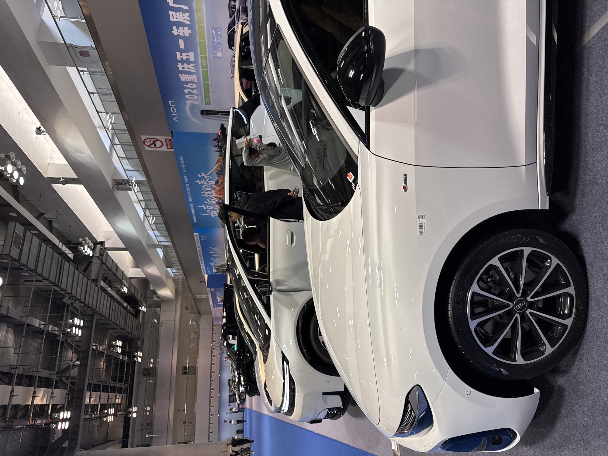 The image captures a car exhibition in Unknown, Unknown, focusing on a row of white Audi cars parked on a blue carpet. The leading car in the foreground showcases its front side, including the tire and rim. Several people are visible inspecting the cars, some inside the vehicles. Banners and advertisements can be seen in the background, including one that reads "2026 重庆五一车展厂" and "ΛΙΟΠ". The exhibition hall is illuminated by overhead lights.