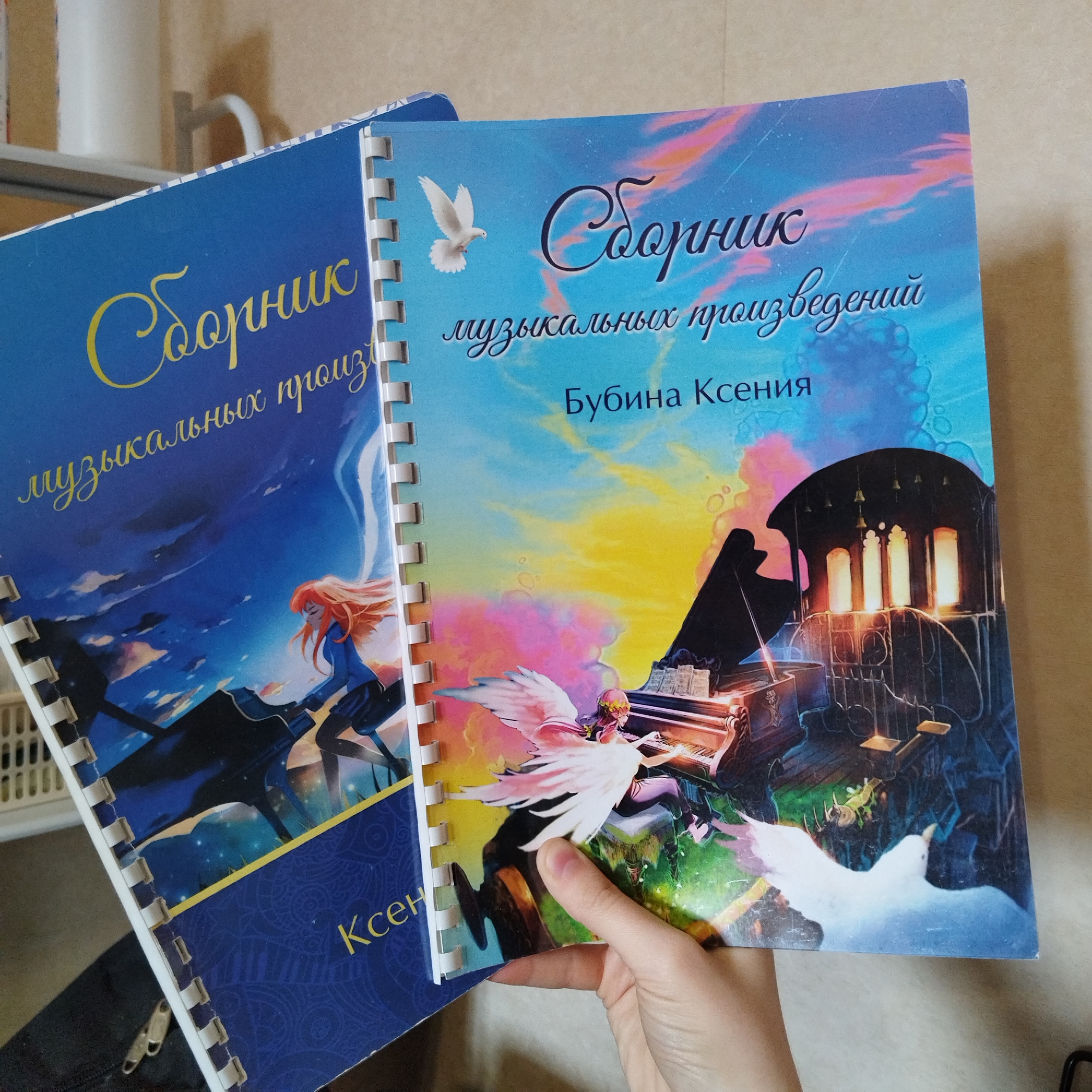 The image shows two spiral-bound books, likely music collections, positioned slightly angled on an unknown surface, with a hand holding the rightmost book.
The book on the left has a dark blue cover with golden yellow cursive text. The visible text reads "Сборник музыкальных произведений" (Collection of musical works) and partially "Ксен" (Ksen). The illustration on its cover depicts a figure with long reddish-orange hair, dressed in a blue outfit, appearing to be interacting with a large black piano or similar musical instrument. The background is a dark, cloudy, and starry sky.
The book on the right is held by a light-skinned hand, showing its full cover. The cover features a vibrant sky transitioning from light blue at the top to yellow, pink, and green hues towards the bottom. White cursive text at the top reads "Сборник музыкальных произведений" (Collection of musical works), and below it, "Бубина Ксения" (Bubina Ksenia) is written in a simpler black font. The illustration includes a white dove flying at the top left. A central figure with large, feathery white and pink wings is depicted playing a grand piano. To the right, a dark, ornate structure with illuminated arched windows resembles a building or part of a cathedral. Another white bird is seen flying near the bottom right.
Both books are bound with white plastic spirals. The overall theme of the covers combines fantasy elements with musical instruments, suggesting a collection of music. The background is indistinct, showing light-colored walls or furniture.
