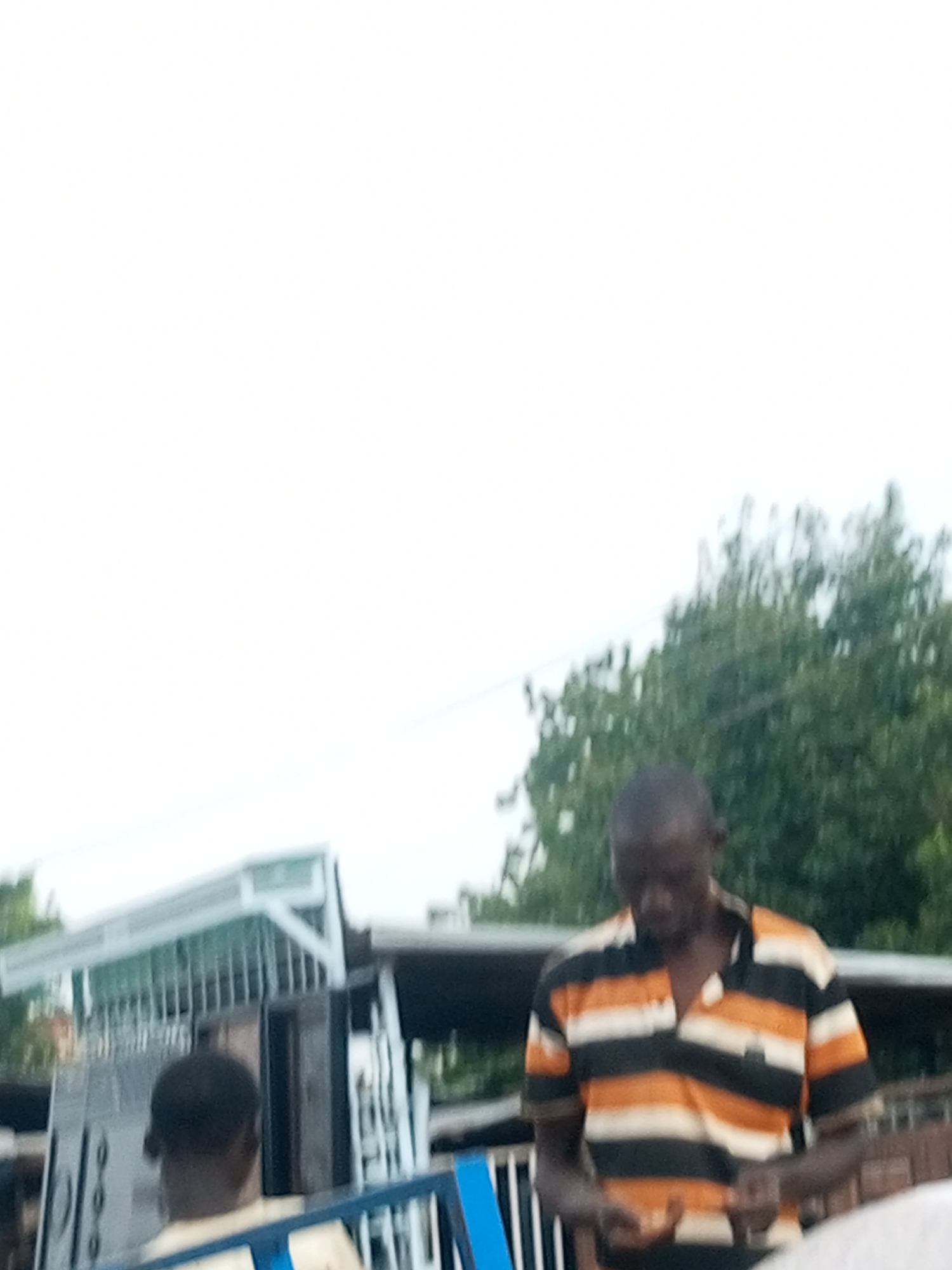 In Garko, Nigeria, on a cloudy day, a Black man is standing outdoors. He is wearing a horizontally striped polo shirt with colors of orange, black, and white. His head is bowed as he looks down at his hands. To his left, the back of another Black person's head is visible, their face turned away from the camera. Behind the man, a metal structure with vertical bars and a white roofed canopy is visible, suggesting it could be a stall or an outdoor workspace. Lush green trees form the background, with the sky appearing overcast and light.
