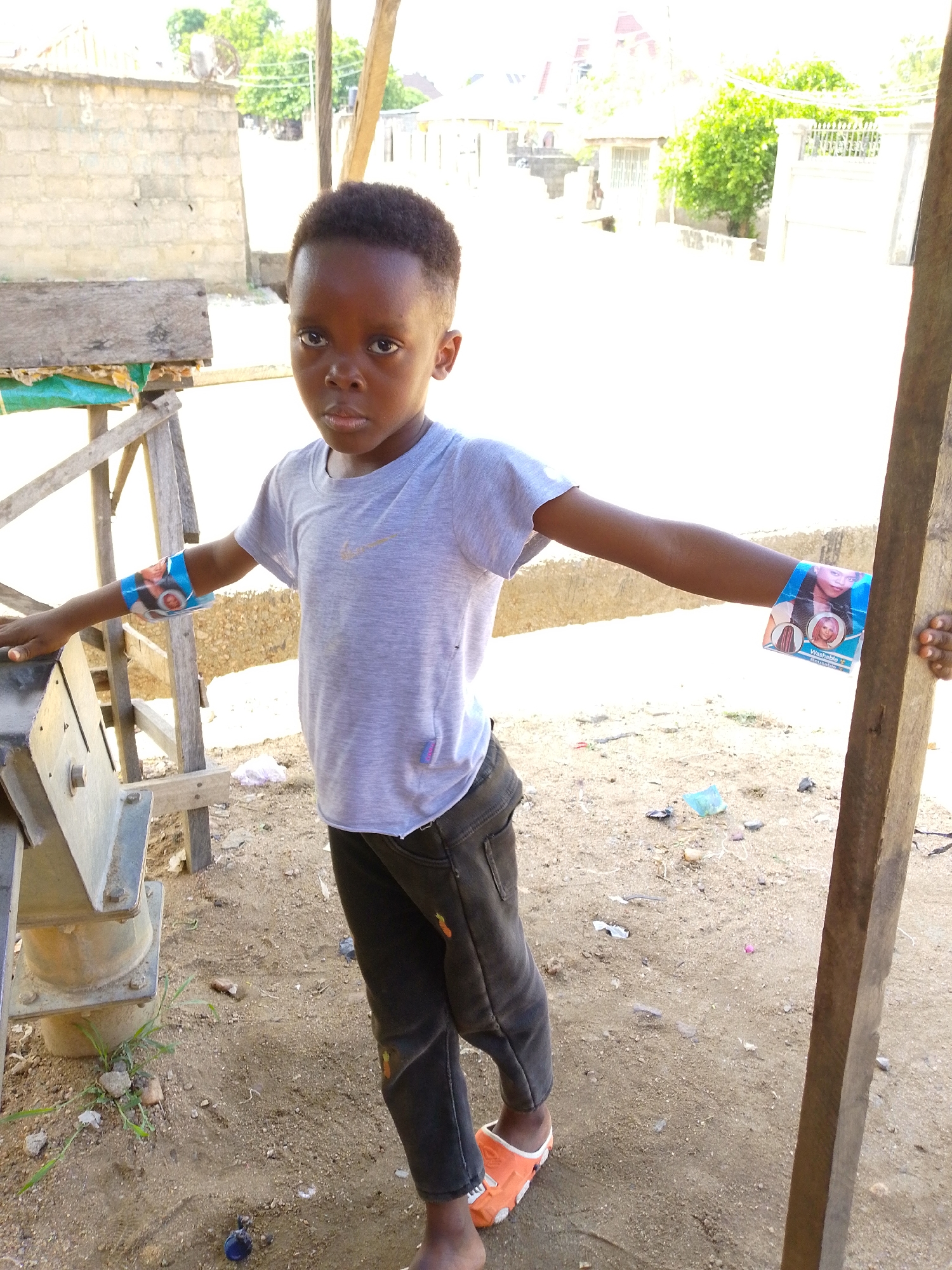 In Lokoja, Nigeria, a young Black child stands outdoors on a dirt surface, their arms outstretched to either side. On each arm, a colorful piece of paper is affixed. The child wears a gray t-shirt, dark pants with a possible decorative element near the lower leg, and orange footwear. Their gaze is directed toward the viewer. The backdrop comprises structures and foliage, with a wooden structure nearby. Litter is scattered on the ground.
