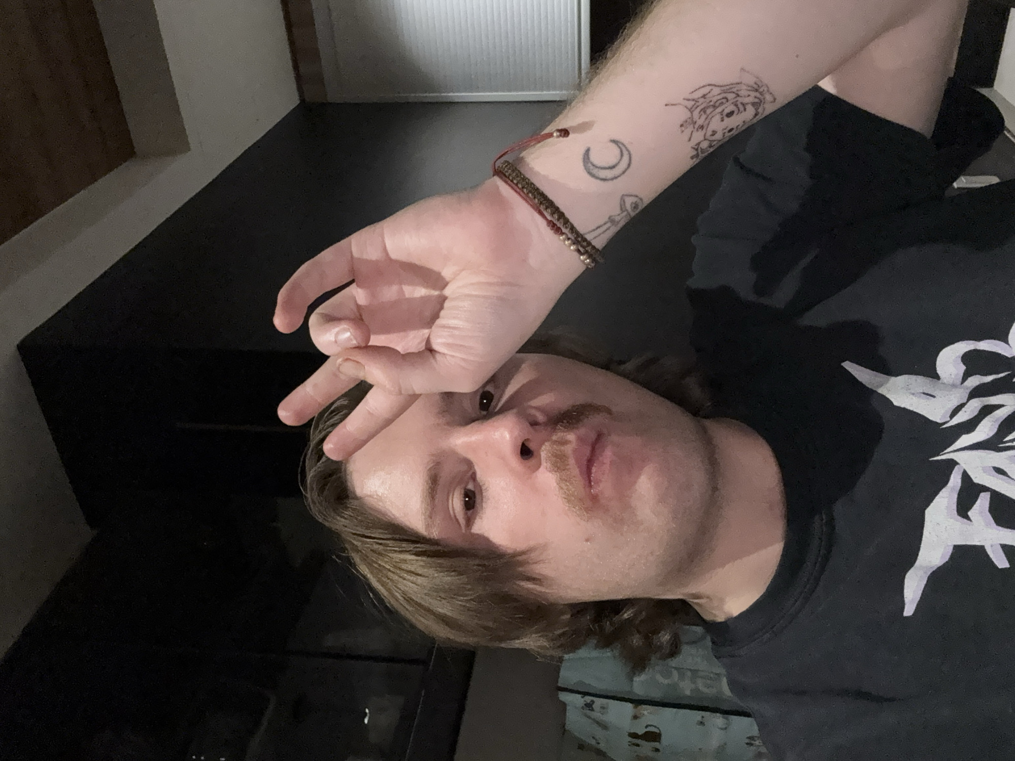 An overhead-angled medium shot captures a person, likely male, with light skin, brown hair, and a mustache, making a peace sign gesture with their right hand. The person's head is tilted, and they gaze towards the camera with a neutral to slightly relaxed expression.
Visible on their right arm are several tattoos, including a crescent moon, a key or similar object, and an abstract design closer to the shoulder. A woven bracelet with small beads is worn on their wrist. The person is wearing a black t-shirt featuring a prominent white, abstract graphic design on the front.
The setting appears to be indoors with subdued lighting, suggesting an evening or low-light environment. The background primarily consists of a dark, reflective surface, possibly a floor, and a lighter wall or partition with faint vertical lines, potentially blinds or paneling. There is no discernible text visible in the image.
Based on the provided context, the image is set in Huixquilucan de Degollado, Mexico, although no specific visual cues within the image confirm this location.