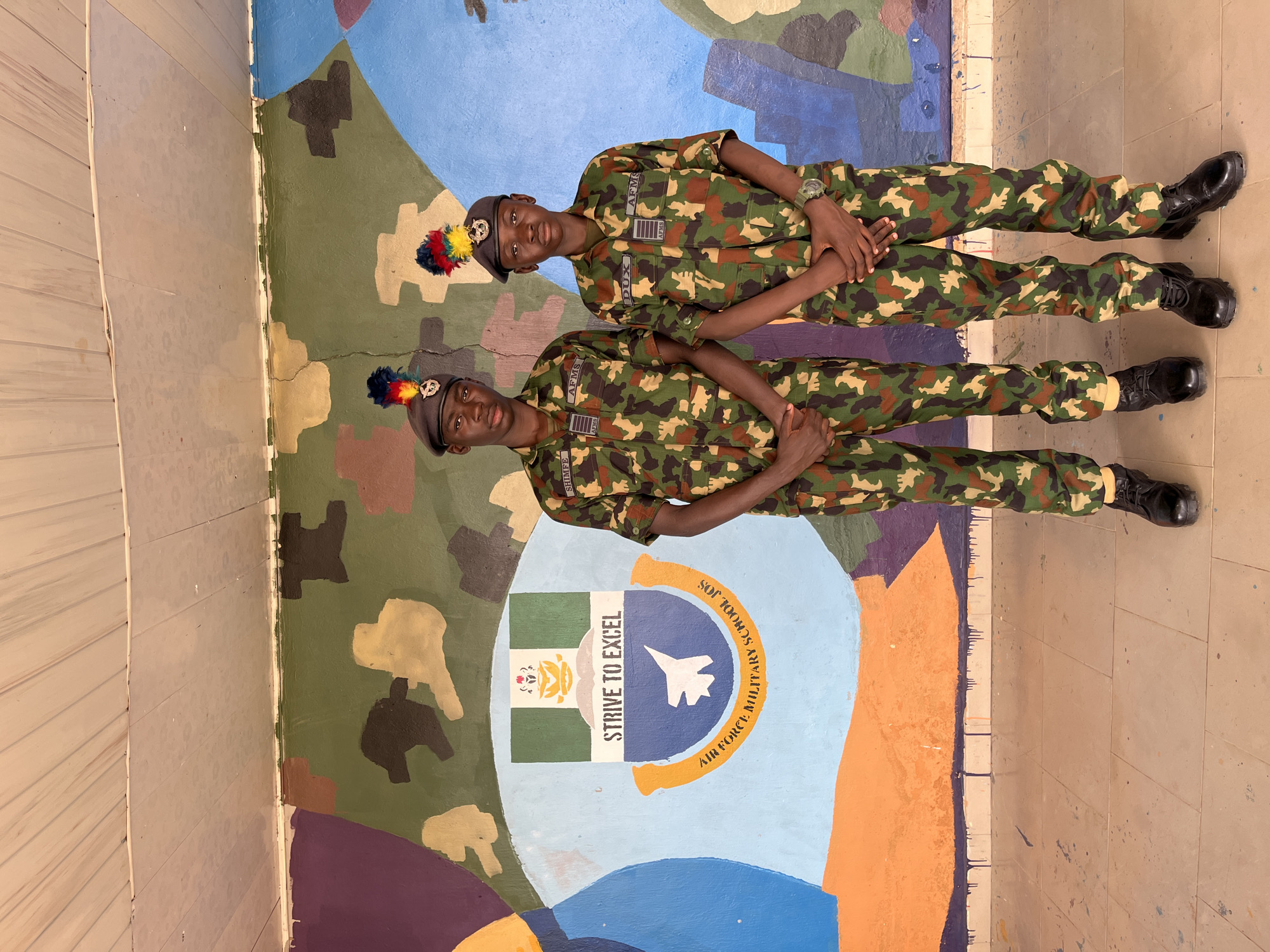Two young males in Nigerian Air Force Military School (AFMS) uniforms stand in front of a camouflage-patterned mural. Both individuals wear camouflage uniforms, dark berets with colorful plumes, and black boots. The mural behind them features the AFMS crest, which includes a Nigerian flag, the motto "STRIVE TO EXCEL," and an image of a fighter jet. The mural is painted on an exterior wall, with a tiled floor in the foreground and what appears to be a wooden ceiling above. The overall scene suggests an outdoor setting at the Air Force Military School in Jos, Nigeria.
