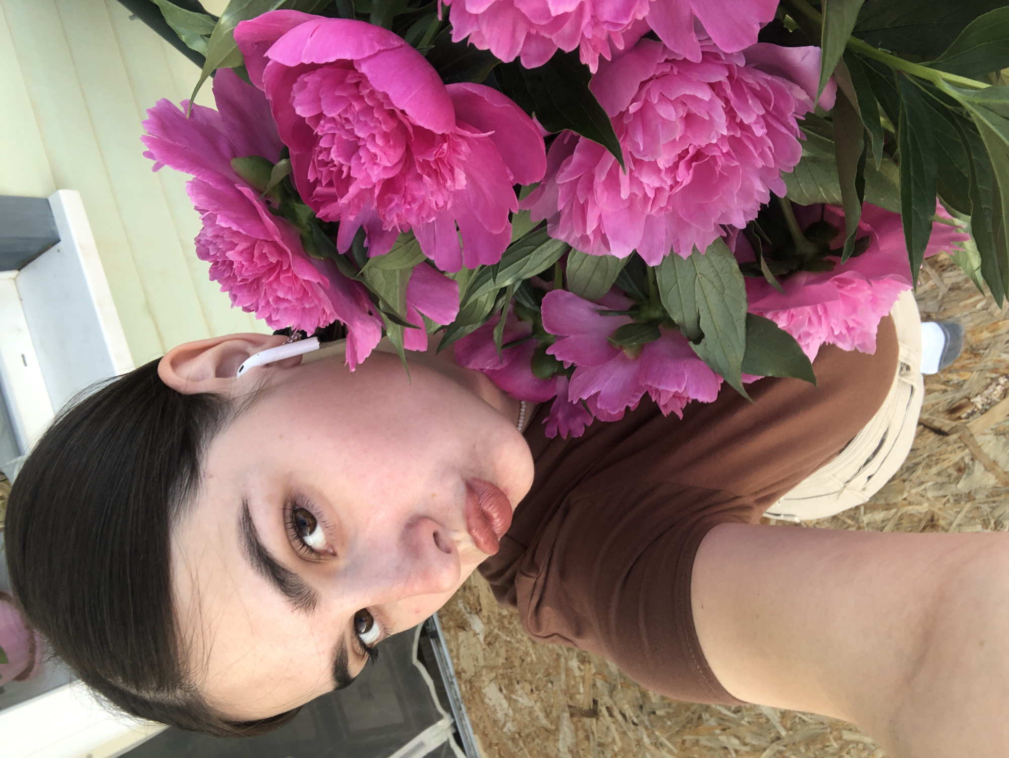In this portrait, a young woman with dark hair and brown eyes is captured from the chest up. She is holding a bouquet of vibrant pink peonies, which partially obscure her face. The flowers are lush and full, with delicate, ruffled petals. She is wearing a brown t-shirt and a light-colored skirt or pants. A white earbud is visible in her right ear. Her expression is playful, with her lips pursed and her head tilted. The background suggests an outdoor setting with a light-colored wooden siding and what appears to be a plywood surface. The lighting is bright, indicating it is daytime. The image is oriented sideways. No text is visible in the image. The location context is Noviy, Russia.