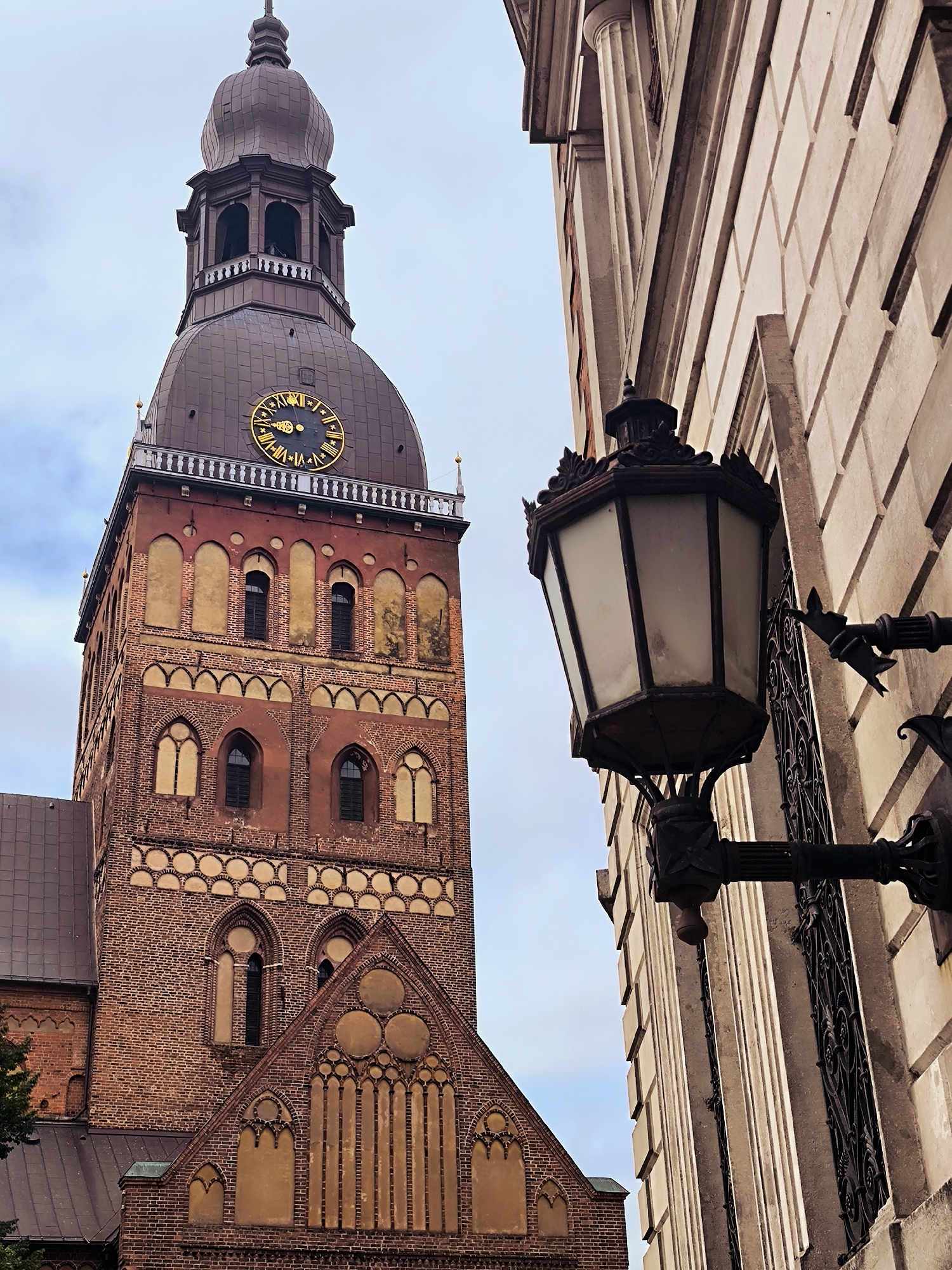 This image shows a portion of the Riga Cathedral in Riga, Latvia, captured from a low angle. The foreground features an ornate, dark metal lantern mounted on a pale stone building to the right. The lantern has a hexagonal, frosted glass enclosure and intricate scrollwork on its arm and mounting. Beyond the lantern and to the left is the imposing brick tower of the Riga Cathedral. The tower is constructed of reddish-brown bricks and features multiple levels with arched windows. A large clock face with Roman numerals is prominently displayed on the tower, just below the copper-domed spire. The sky is overcast and pale blue, with hints of clouds. The architectural style suggests a historical European setting.