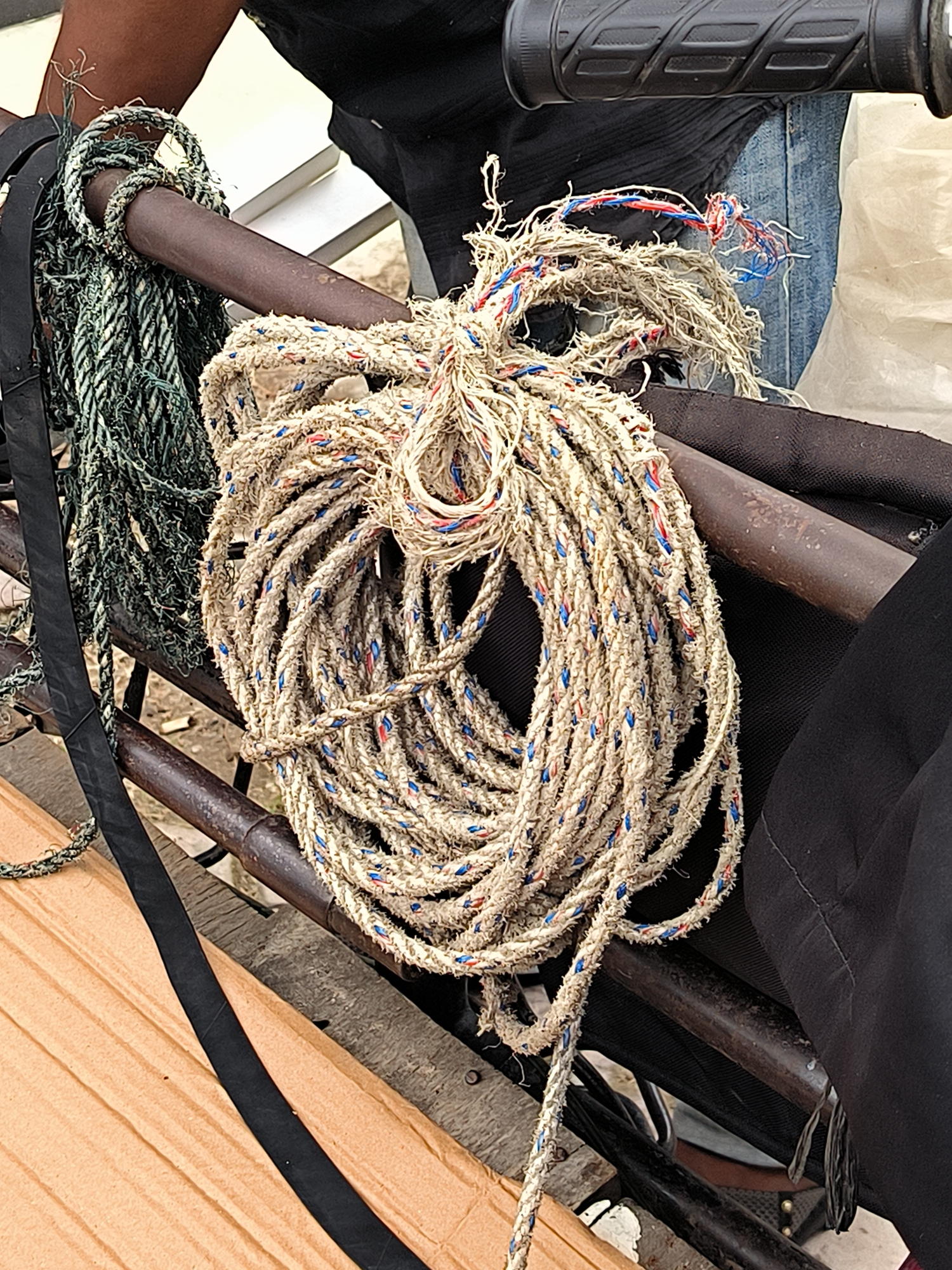 A close-up, slightly elevated, medium shot focuses on two coiled ropes, one thick and beige with red and blue flecks, the other thinner and dark green, resting on a dark metal frame. The beige rope is in the foreground, its loose strands frayed at the top, with a knot of red and blue threads visible. The green rope is behind and to the left, also coiled. A dark brown metal bar runs horizontally across the frame, supporting the ropes. Behind the ropes, a person's dark-clothed torso and arm are partially visible, along with portions of blue denim jeans and a white plastic sheet. In the lower left, a section of light brown corrugated cardboard is present. The overall impression is of a utilitarian scene, possibly in a workshop or storage area, with a focus on the texture and condition of the ropes, suggesting wear and use. The scene is static, with no discernible activity or interaction. The location is Lhokseumawe, Indonesia.
