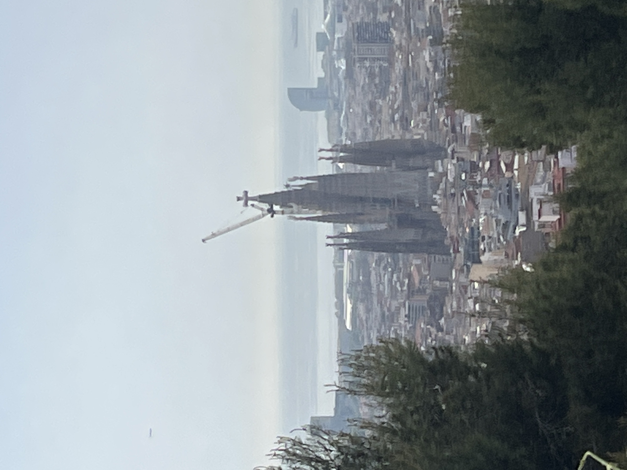 The image displays a panoramic, high-angle view of Barcelona, Spain, under an overcast or hazy sky, viewed from an elevated position. In the mid-ground, the iconic Sagrada Familia cathedral stands prominently on the left, recognizable by its distinct spires and an attached construction crane, indicating ongoing building work. The dense urban landscape of Barcelona fills the scene, with numerous buildings extending towards the distant coast.
To the right, lush green trees and foliage occupy the foreground, suggesting the photograph was taken from a hill or park overlooking the city. The Mediterranean Sea forms the horizon, appearing light and somewhat indistinct due to atmospheric conditions. A distinctive curved building, possibly the W Hotel Barcelona, is visible near the coastline in the background, further confirming the location.
The time of day appears to be daytime, characterized by diffused light and a lack of sharp shadows. No specific activities or events are depicted beyond the perpetual construction of the Sagrada Familia. There are no visible people or discernible text within the frame.