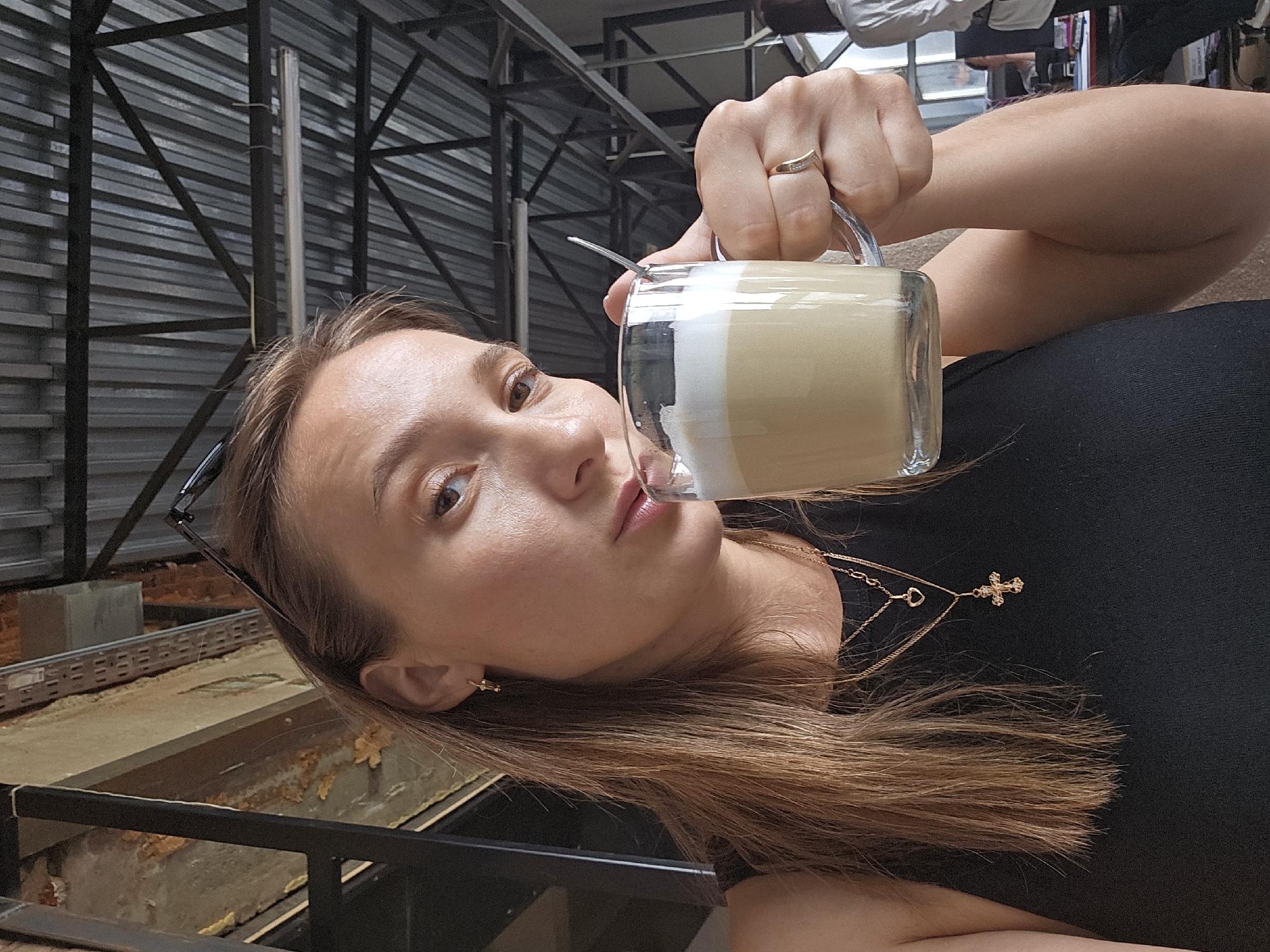 A fair-skinned woman with long brown hair and a gold cross necklace is holding a glass mug filled with a beverage that appears to be a latte or similar milk-based drink. She is wearing a gold ring on her right hand. Her head is tilted, suggesting she might be taking a selfie while drinking. Her sunglasses are tucked into the hair. The background features an industrial-looking metal structure and what appears to be the edge of an outdoor seating area. The photograph was taken in the city of Chaikovsky, Russia.