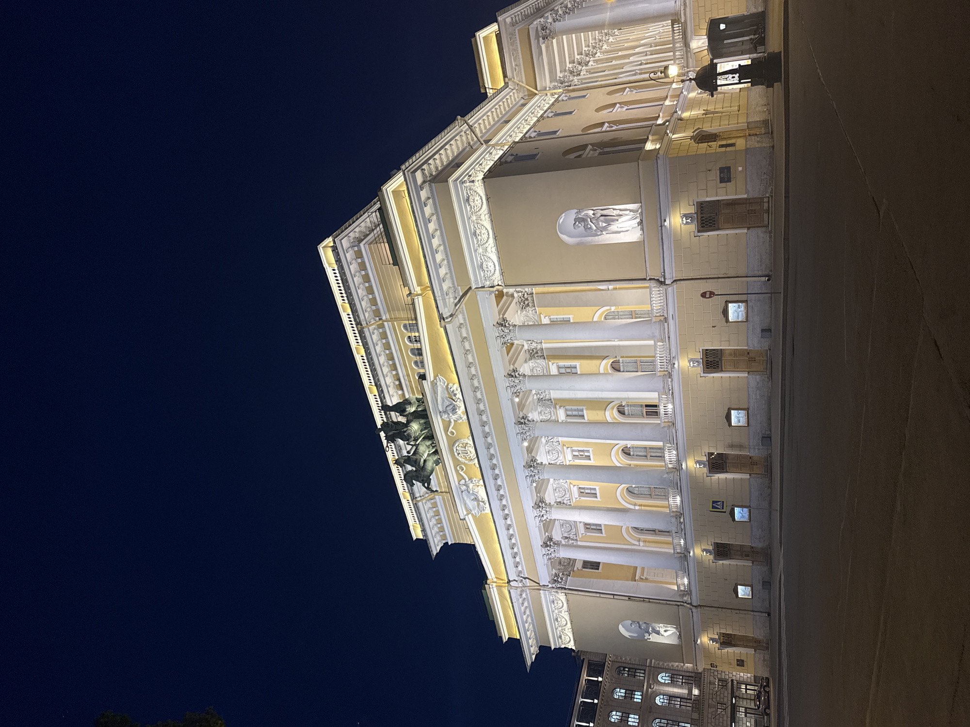 The image captures the facade of the Alexandrinsky Theatre in Saint Petersburg, Russia, at night. The theatre, illuminated by warm lights, stands grandly with its neoclassical architecture. A prominent feature is the Quadriga of Apollo, a sculpture of Apollo driving a chariot pulled by four horses, adorning the roof. The facade is characterized by a series of tall, white Corinthian columns supporting an ornate entablature. Statues are visible in niches along the building's exterior. The street in front of the theatre is paved, and a few streetlights are visible, including an antique-style lamp to the right. The sky is a deep, dark blue, suggesting nighttime. A few cars are parked or driving on the street in the distance. The overall scene conveys a sense of historical grandeur and nocturnal stillness.