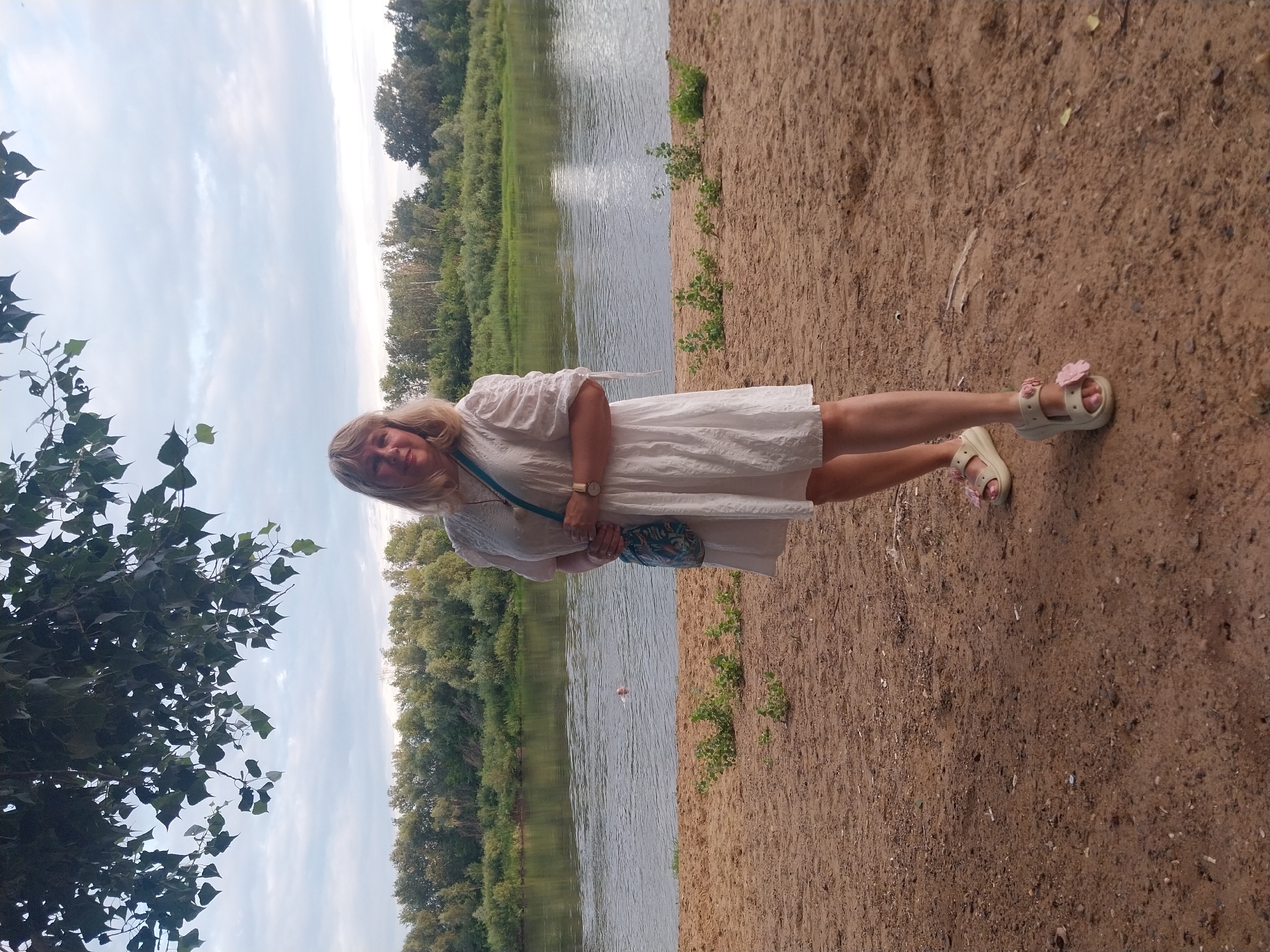 A woman with blonde hair, wearing a white dress and sandals, stands on a sandy shore. Behind her, a wide river flows, and on the opposite bank, a dense green forest extends. The sky above is overcast with clouds. In the distance, a person is swimming in the river. The overall scene is a natural landscape with a person posing for a photograph. There are no identifiable landmarks or signs that would place this image specifically in the city of Orenburg, Russia.