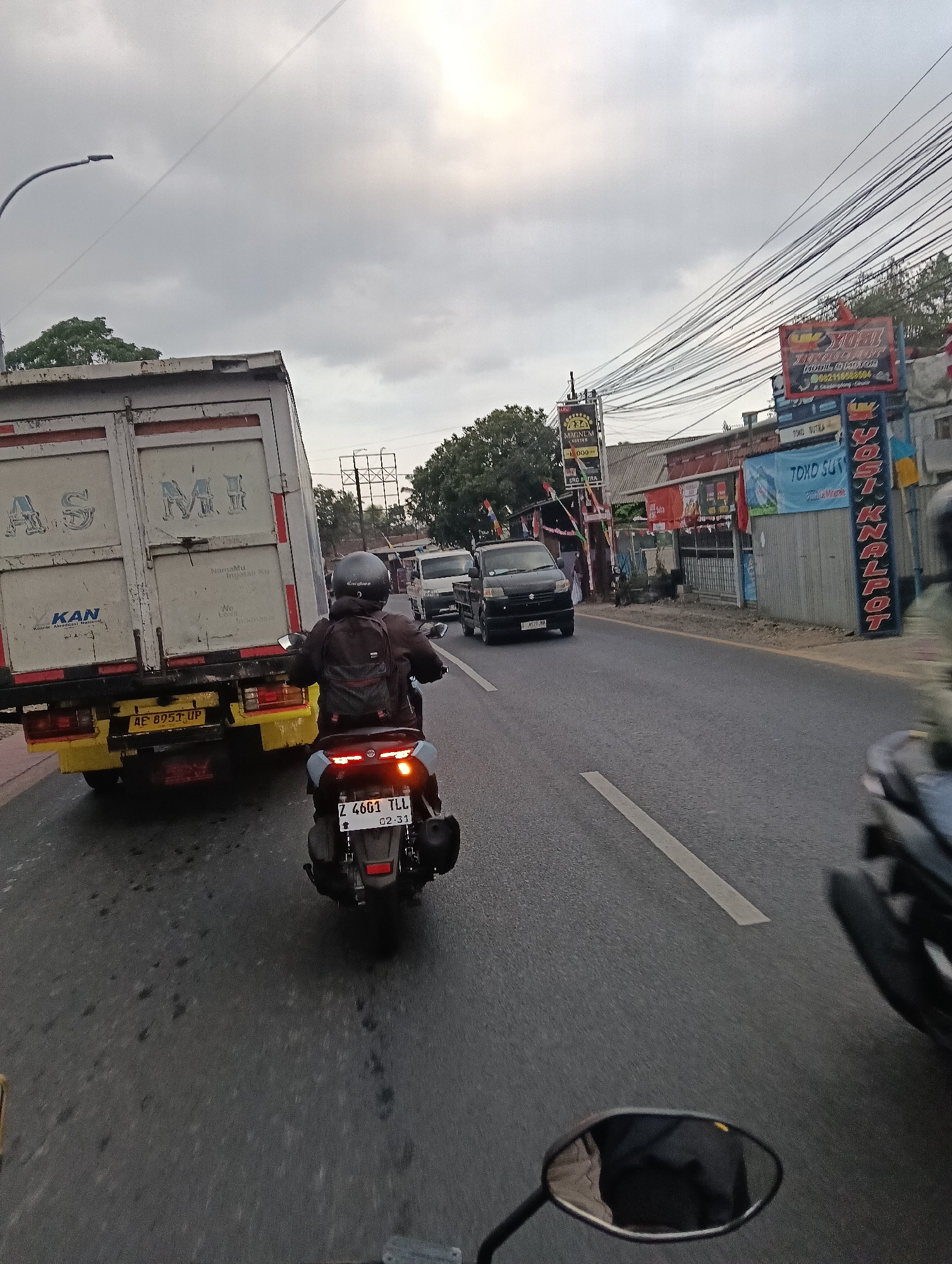 This is a street-level, vertical, eye-level, full shot of a road with traffic in the city of Linggajaya, Indonesia. The scene is captured on an overcast day, with a gray sky dominating the upper portion of the frame. A large, white and yellow box truck with "AS MI" and "KAN" written on its side is on the left, taking up a significant portion of the image. A black motorcycle with a rider wearing a helmet and backpack is in the foreground, moving in the same direction as the truck. The motorcycle's license plate is visible, reading "Z 4661 TLL 02.31". In the background, there are several buildings and shops lining the road, some with signs. One sign in red, white, and blue reads "YOSI KNALPOT". Another sign is for "TOKO SUTRA". A few other vehicles, including a black minivan with license plate "B 8926 NH", are also visible on the road. The road itself is asphalt, with a white line marking the lane. Overhead, a dense network of electrical wires crisscrosses the sky. The overall impression is a busy street scene in an Indonesian urban environment.