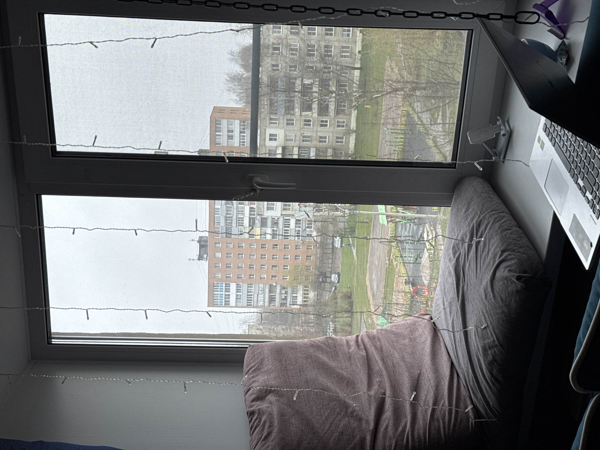 The image presents an interior view looking out through a modern, white PVC window into a residential urban landscape in Ново-Никольское, Russia. Decorative string lights are draped across the window frame and panes, creating a soft visual element.
Inside, to the right of the window, a silver laptop with a visible keyboard and a white phone stand are positioned on a windowsill or desk. A purple clip or hanger and a dark chain are also present near the top right of the frame. On the left side, two large, soft pillows – one grey and one brownish-mauve – are placed against the window, suggesting a comfortable seating or lounging space.
The exterior view reveals several multi-story residential buildings, typical of post-Soviet architecture, with numerous windows. Bare trees and patches of green grass are visible between the structures, along with paved areas that might be courtyards or paths. The sky is uniformly overcast and grey, indicating a dull, possibly early spring or late autumn day. No individuals are visible. The laptop's keyboard shows standard English QWERTY characters, but no specific text is legible. The scene conveys a quiet, possibly contemplative atmosphere from an indoor vantage point.