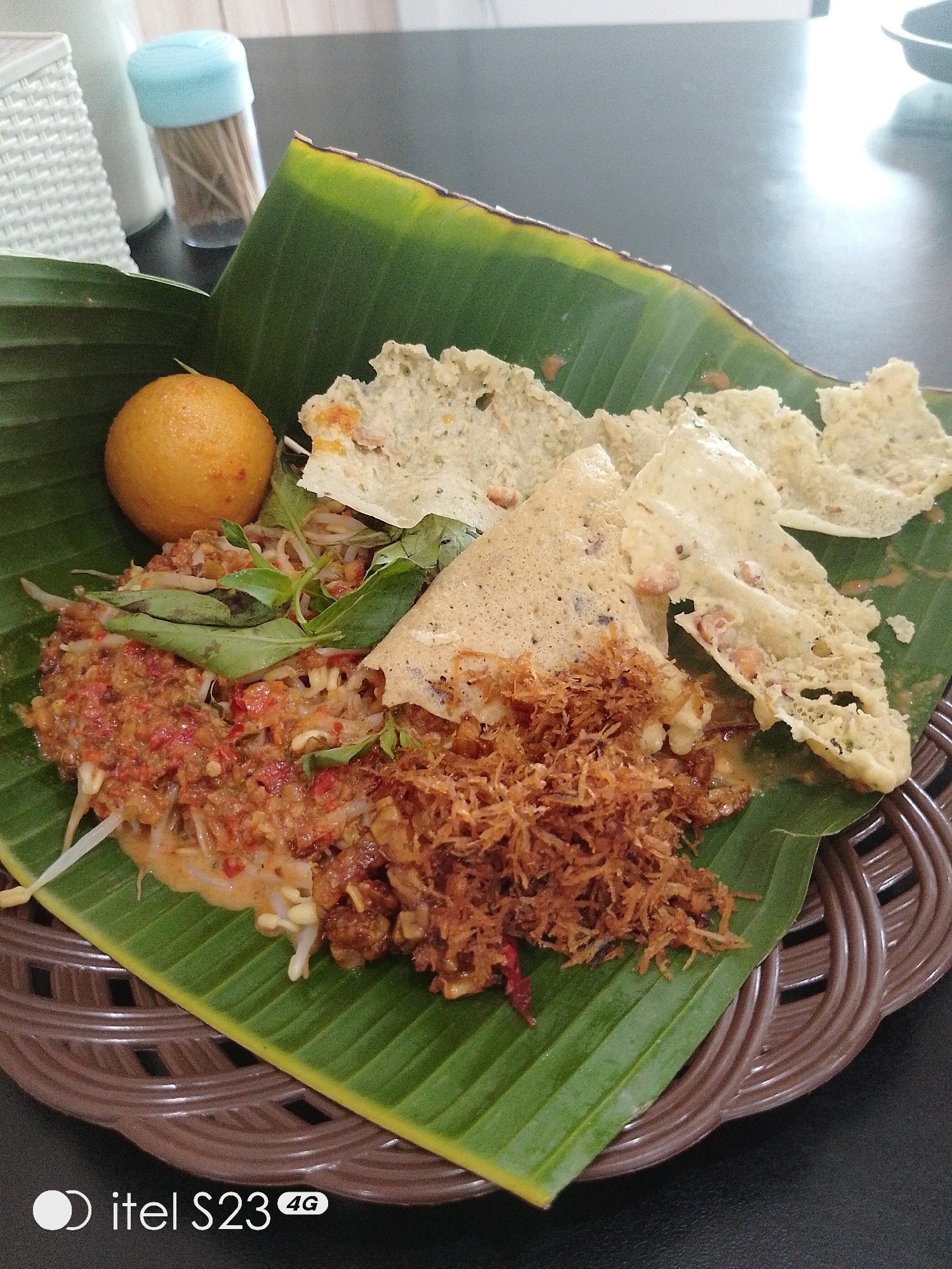 A close-up, slightly overhead shot captures a portion of a traditional Indonesian dish served on a woven brown plate lined with a vibrant green banana leaf. The dish appears to be a type of mixed vegetable salad with a reddish sauce, possibly rujak. It is topped with shredded coconut, sprouts, and fresh green herbs. To the left of the salad, a whole, round, orange-colored item, possibly a boiled egg or a piece of fruit, sits on the banana leaf. Several pieces of light-colored, crispy crackers, resembling krupuk, are scattered around the dish. In the blurred background, a container of toothpicks with a light blue lid is visible, along with a white, woven object. The lighting suggests an indoor setting, possibly a restaurant. The bottom left corner of the image features a watermark that reads "itel S23 4G". The overall impression is one of a colorful and appetizing culinary presentation. The dish is commonly found in Indonesian street food stalls and local eateries, including those in the city of Surabaya, Indonesia, where such authentic culinary experiences are prevalent.