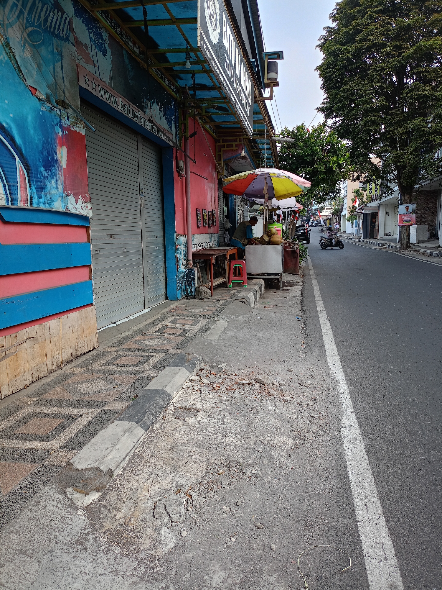 The image depicts a street scene in Batu, Indonesia, on a bright day. In the foreground, a sidewalk is visible, paved with a patterned tile design and bordered by a gray curb. To the left, the facade of a building is adorned with blue and red horizontal stripes and a large banner with blue and white graphics. A gray roller shutter door is visible on this building.
Adjacent to the building and sidewalk, a street vendor is operating from a cart, under a colorful umbrella with yellow, red, and green sections. The vendor, a man wearing a blue shirt and vest, appears to be preparing coconuts, with several whole coconuts and cut pieces visible on the cart. A red stool is placed near the cart.
The street itself is paved with asphalt and marked with a white line. Traffic is light, with a single motorcycle visible in the middle distance. Buildings line the opposite side of the street, some with awnings and signage, and a large tree with dense foliage stands on the right side of the image. The sky is bright and clear, suggesting fair weather. The overall impression is of a typical urban commercial street.