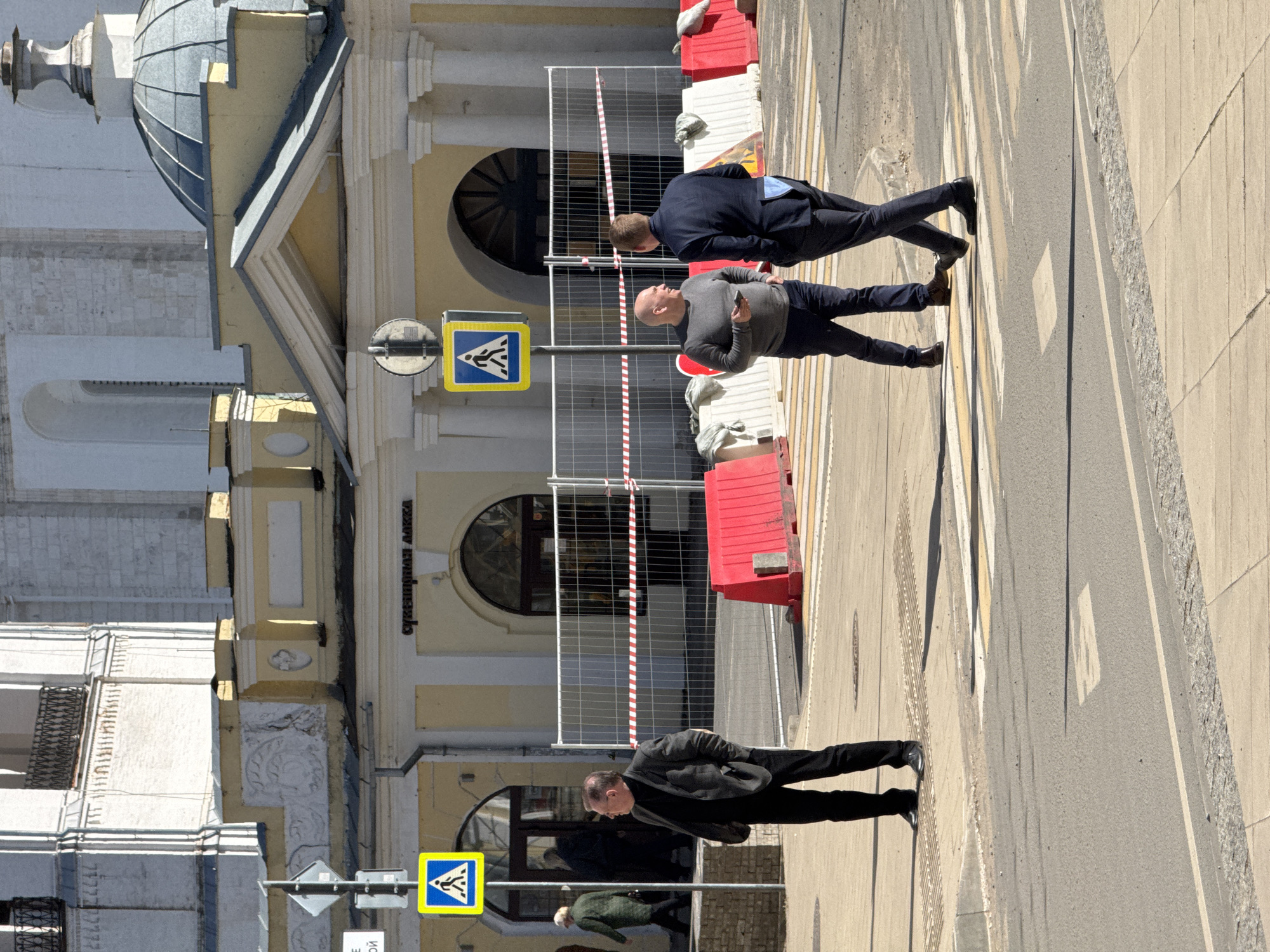 The image depicts a street scene in Novo-Nikol'skoe, Russia. Three men are visible in the foreground: two are crossing the street at a pedestrian crosswalk, and one is walking on the sidewalk. A crosswalk sign is present. Temporary fencing and red barriers are set up near the crosswalk, suggesting nearby construction. A yellow building with architectural detailing, including a dome, is situated beside the street.
