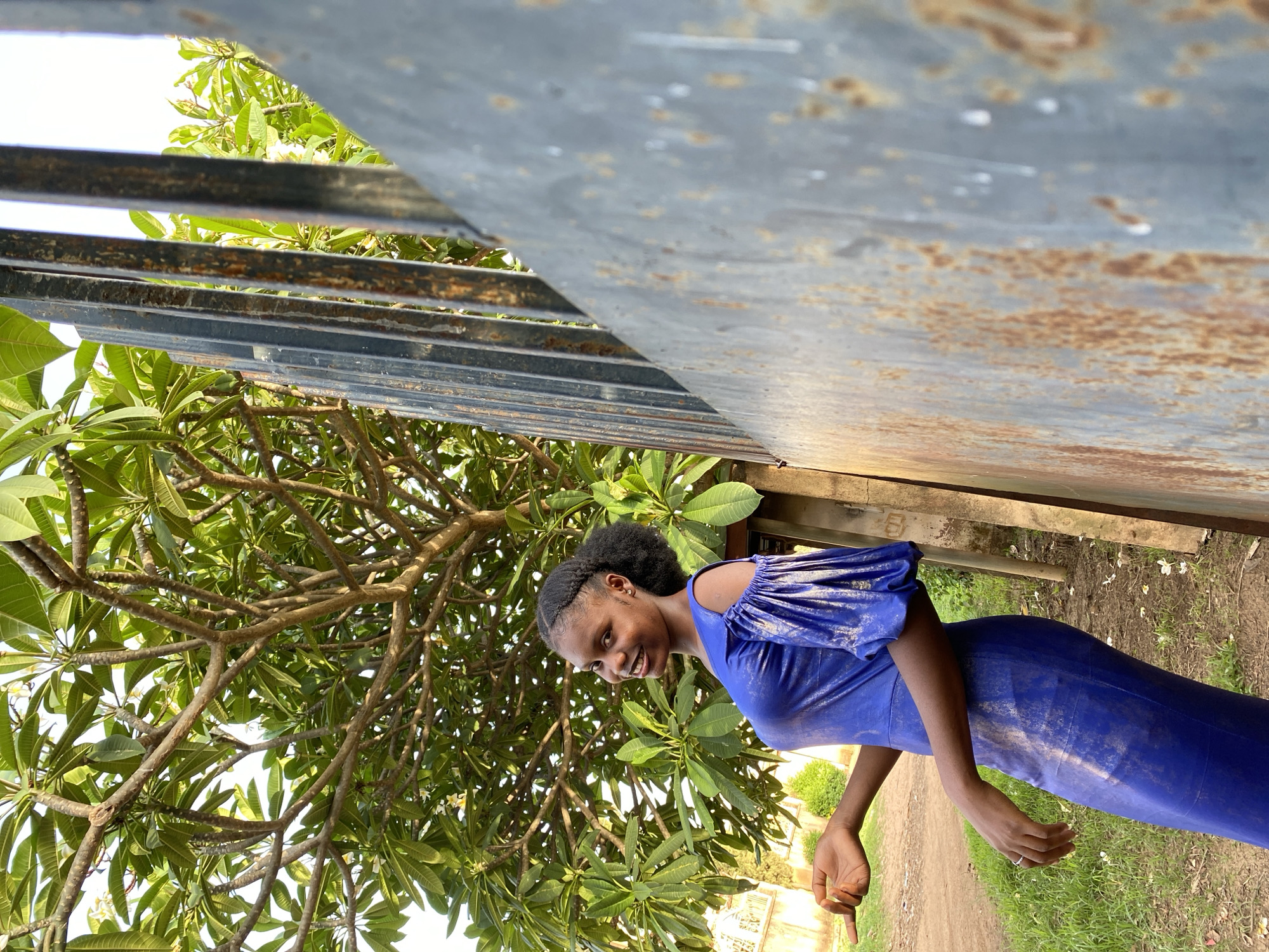 A young woman in a blue, fitted dress stands near a rusted metal structure and a large tree with green leaves. The location is Vom, Nigeria, indicated by the background vegetation and building style. The woman is smiling and appears to be pointing or gesturing. She is standing on a dirt path with grass. The metal structure is overhead and to the side, showing signs of corrosion. A tree branch with large leaves is visible in the foreground.