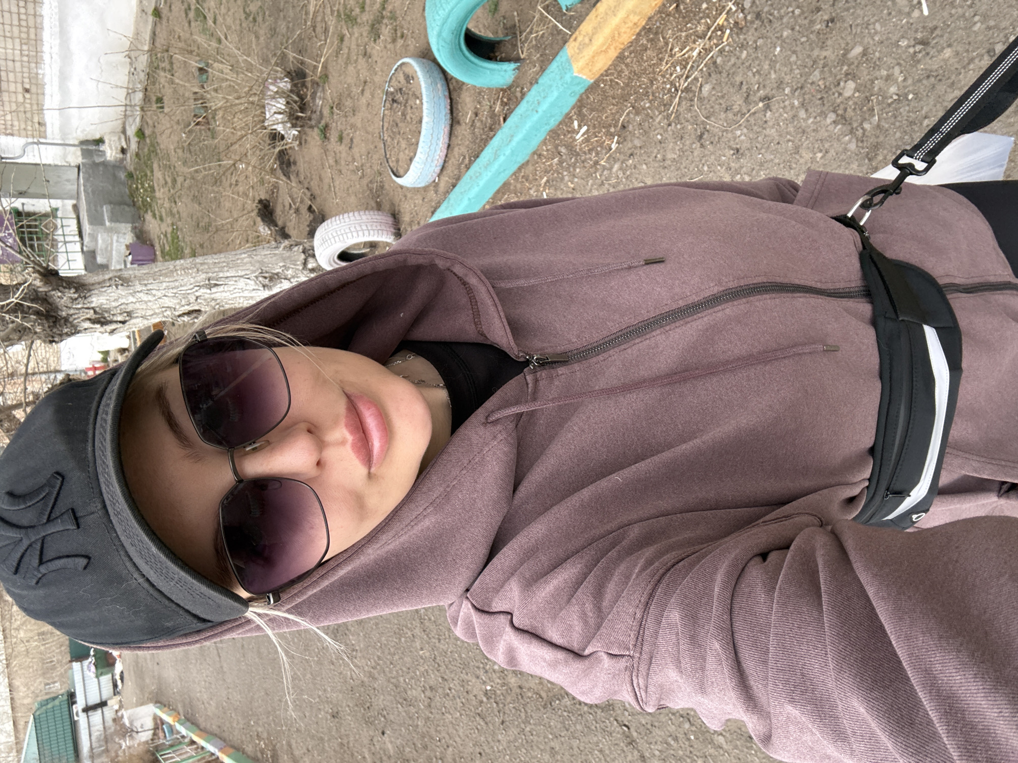 A woman with fair skin is centered in an outdoor selfie. She wears a dark baseball cap with a light-colored logo, dark sunglasses, and a mauve-colored hooded jacket with a partially zipped front. A black fanny pack with a white stripe is visible across her torso. The background shows an urban landscape. In the upper portion of the image, three painted car tires lie near a painted metal beam. Bare trees and dry ground are also visible. The lower portion shows a small, green and brown metal fence. The location is speculated to be Borzya, Russia.