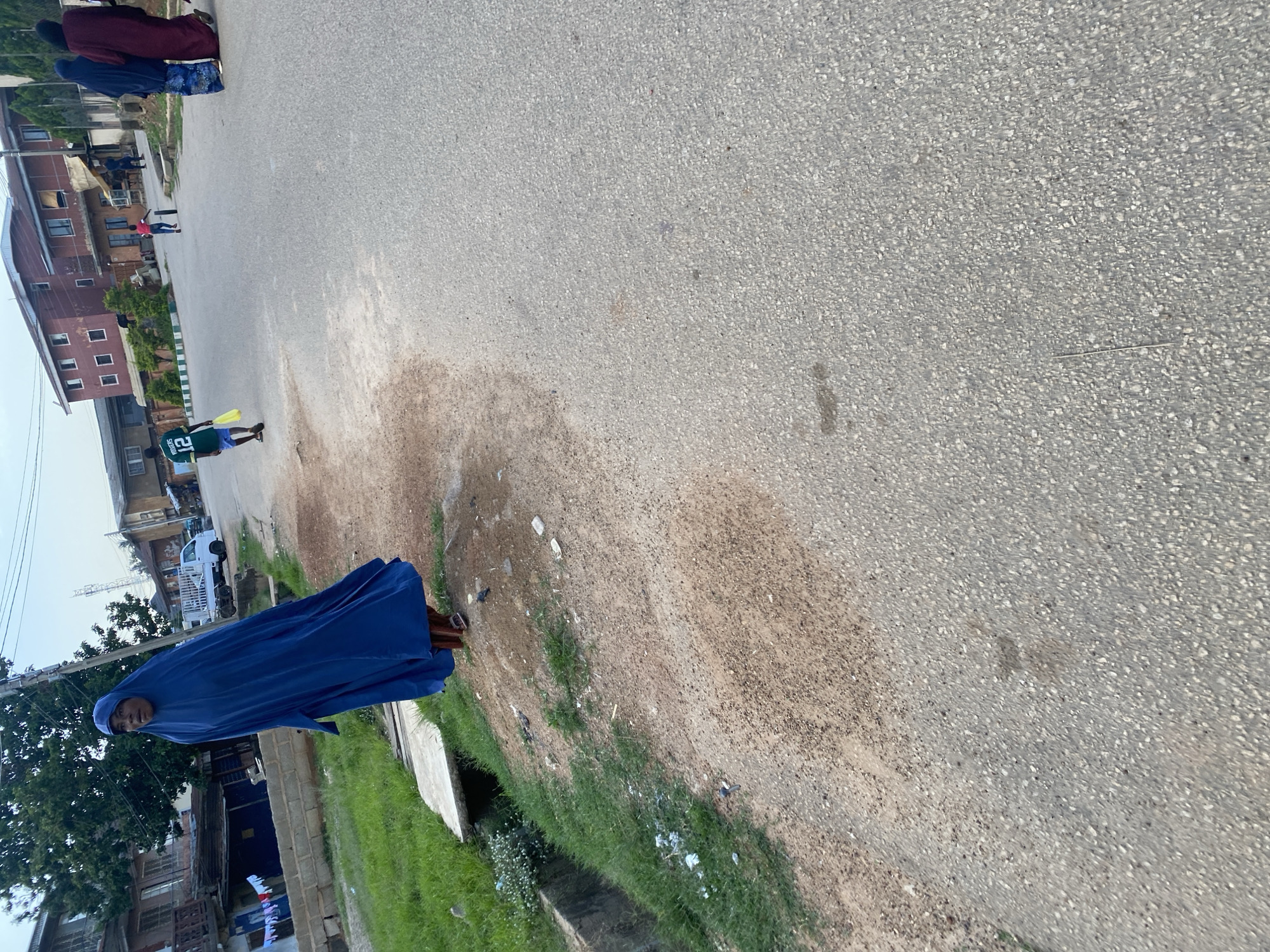 The image depicts a street scene in Jos, Nigeria. In the foreground, a person dressed in a long, flowing royal blue hijab stands on the right side of a paved road. Their feet are visible, wearing brown sandals. To the left of the road, there is a ditch with green grass growing alongside it, and some scattered debris. Further down the road, a young person in a green and white jersey with the number 12 is walking away from the camera, carrying a yellow plastic bag. In the background, several buildings are visible, including a multi-story red building and other structures that appear to be houses or shops. A white truck is parked on the side of the road. Two individuals in traditional Islamic attire, one in dark blue and the other in maroon, are walking away from the camera on the right side of the road. The sky is overcast.
