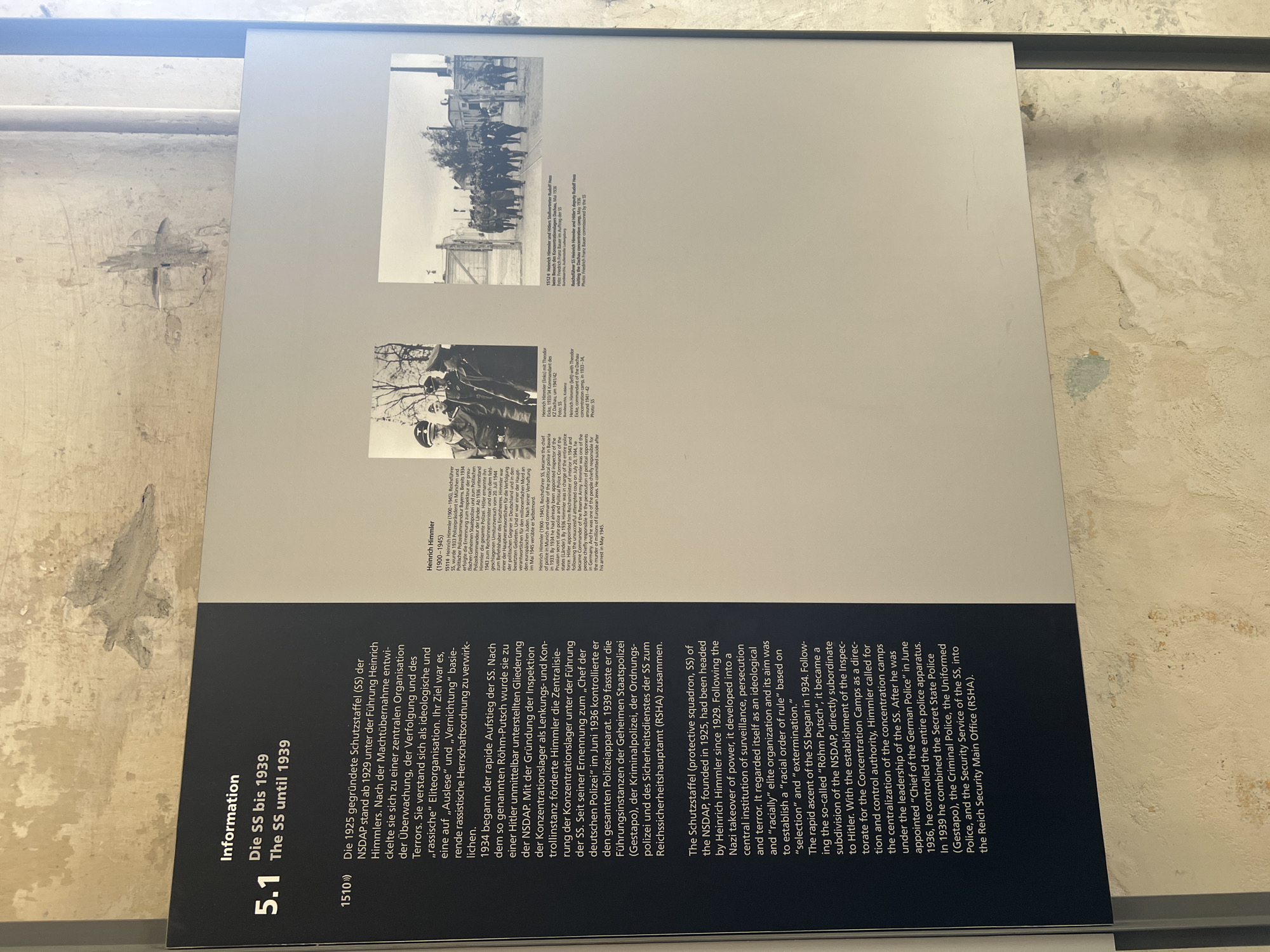 The image shows a display panel inside a museum, likely at the Dachau Concentration Camp Memorial Site, given the context of the text and images. The display has a heading "Information" and a section titled "5.1 Die SS bis 1939 / The SS until 1939." The panel includes text in German and English describing the rise of the SS from 1925 to 1939, its leadership under Heinrich Himmler, and its transformation into an organization focused on surveillance, persecution, and control, leading to the establishment of concentration camps.
To the right of the text, there are two black and white photographs. The first photograph, captioned "Heinrich Himmler (Links) mit Theodor Eicke, 1933/34 Kommandant des Konzentrationslagers in Dachau, Mai 1936", shows Heinrich Himmler on the left, wearing a uniform with a cap, smiling. Beside him are two other men in similar uniforms, one of whom is identified as Theodor Eicke. The background appears to be an outdoor setting with some trees.
The second photograph, captioned "1932/3 Heinrich Himmler und Hitler's deputy Rudolf Hess visiting the Dachau concentration camp, May 1936. Foto: Friedrich Franz Bauer commissioned by the SS", shows a group of men, presumably prisoners, walking in a line towards a gate. Heinrich Himmler and Rudolf Hess are also present, observing the prisoners. The architecture in the background includes buildings and fences, consistent with a concentration camp setting. The overall environment depicted in the photographs is stark and somber. The display panel itself is mounted on a rough, textured wall, possibly the original structure of a building at the memorial site.