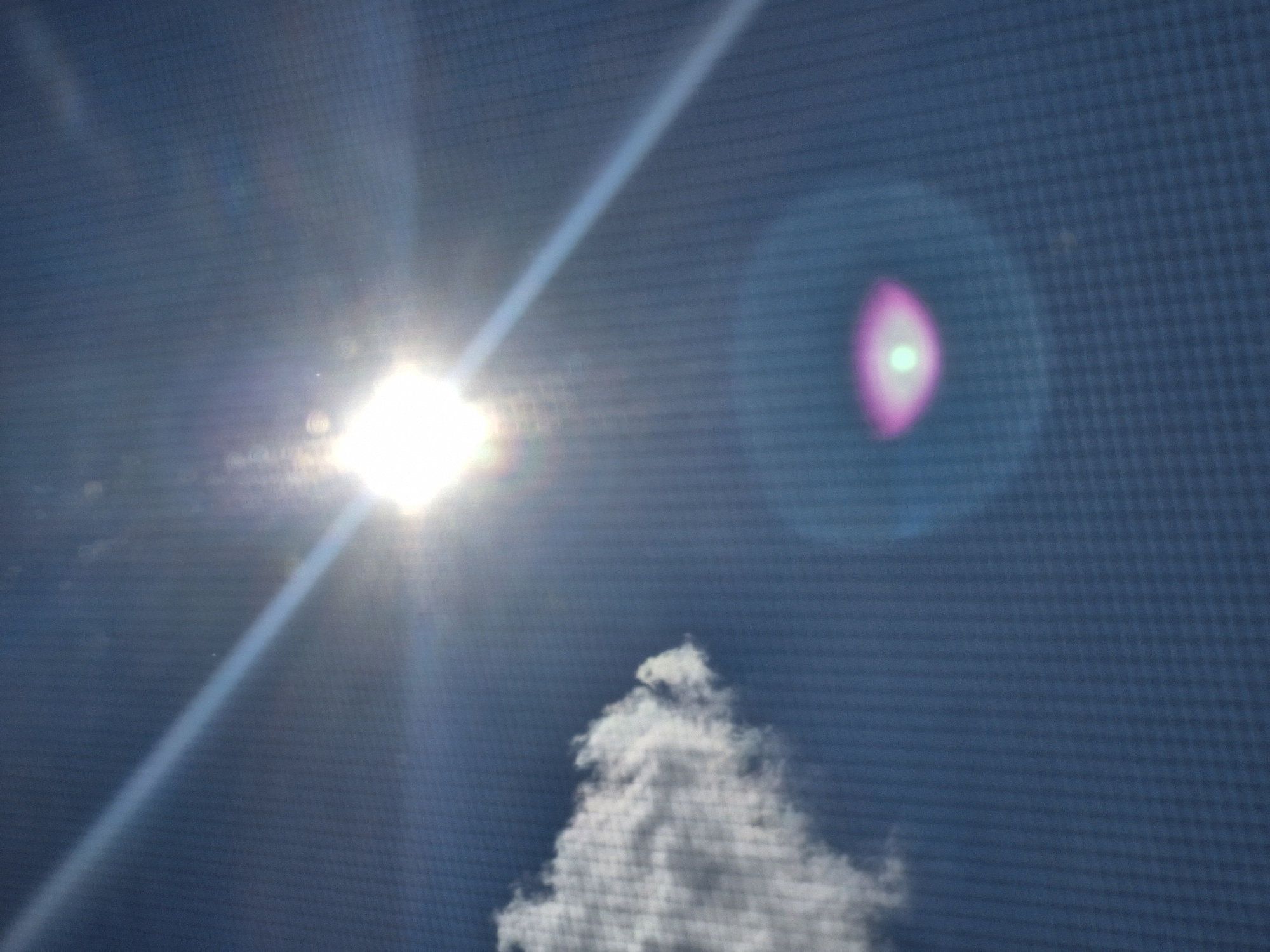 The image depicts a bright sun with a lens flare across a blue sky. A white cloud is visible on the left side of the frame. A circular light artifact with pink and green hues is present in the lower right portion of the image. The image quality is grainy, suggesting it may have been captured through a screen or with a low-resolution camera. There are no people, structures, or identifying features of Kazan, Russia, visible in the image.