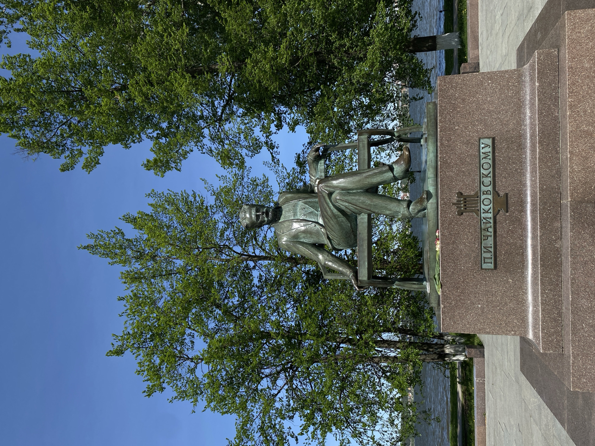 The image depicts the monument to Pyotr Ilyich Tchaikovsky in Votkinsk, Russia. The bronze statue portrays the composer seated on a bench. The monument stands on a multi-tiered pedestal, with the name "П.И. ЧАЙКОВСКОМУ" inscribed vertically. Trees and blue sky are in the background. The Volga river is in the background.