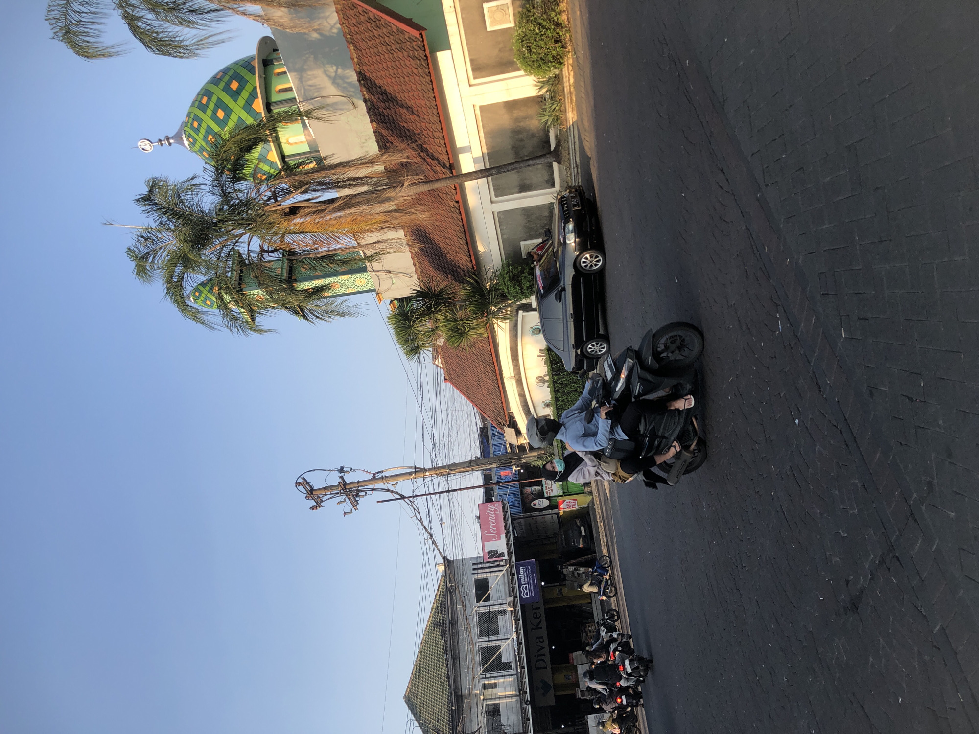 The image captures a street scene in Boro, Indonesia. In the foreground, two individuals are riding a motorcycle. The rider wears a light blue long-sleeved shirt and a helmet, while the passenger, a woman, has a light-colored headscarf and a darker jacket. They are on a black motorcycle with an exhaust pipe visible.
To the right of the motorcycle, a black sedan is parked on the side of the road. Further in the background, a mosque with green, diamond-patterned domes and minarets is visible, with palm trees in front of it. Several other motorcycles are seen on the road, some carrying multiple passengers. Buildings line the street, including a white building with a red-tiled roof and another building with a green tiled roof. A utility pole with numerous wires stands prominently on the left side of the image, near the buildings. The sky is clear and blue, indicating a daytime scene, likely late afternoon due to the quality of light. The road surface is paved with dark interlocking bricks.