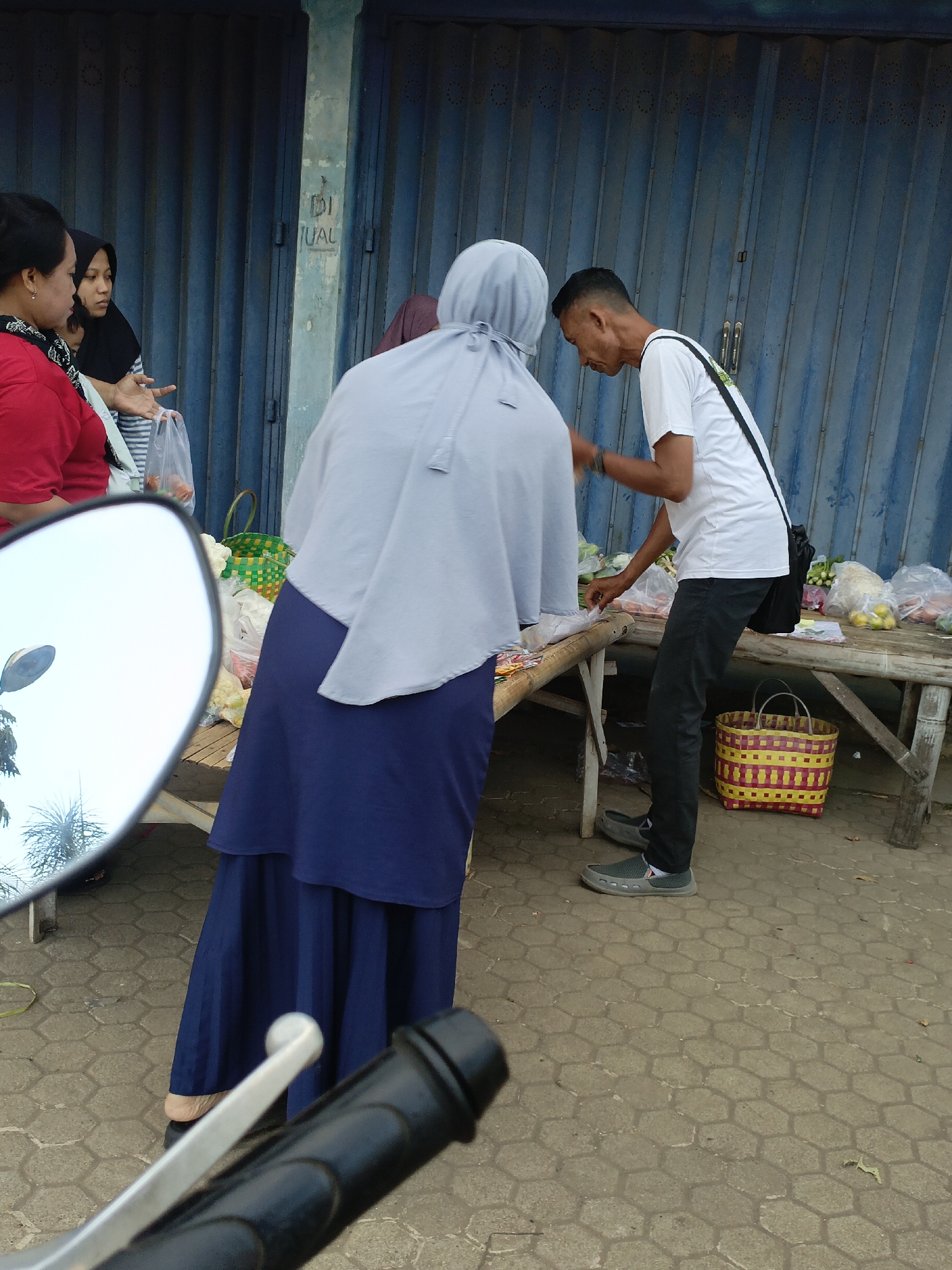 In Srono, Indonesia, at a street-side market, several individuals are engaged in commerce. A woman in a red shirt and a woman in a hijab are at the left of the frame, appearing to be in conversation. The woman in the red shirt is holding a plastic bag. To their right, a man in a white t-shirt and dark pants is attending to a stall laden with produce, including vegetables and what appear to be citrus fruits, arranged on a rustic wooden table. He is interacting with a woman wearing a light blue hijab and a dark blue skirt, who is partially visible. In the foreground, a motorcycle's handlebar and a side mirror are visible, suggesting the photo was taken from a motorcycle. The market is situated in front of a blue, corrugated metal shutter. A colorful woven basket sits on the ground near the stall. The ground is paved with hexagonal concrete tiles.
