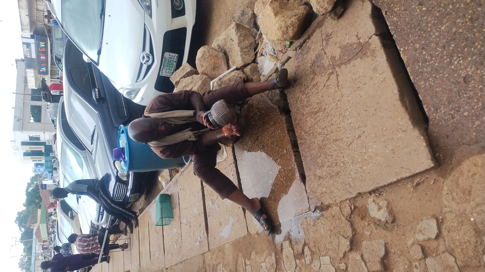 The image captures an outdoor urban scene, rotated 90 degrees clockwise. In the foreground, a man in a brown tunic and trousers, with a light-colored shawl, is squatting and pouring water from a striped pitcher over his hands. He appears to be performing ablution or washing, with water splashing and pooling on the cracked concrete pavement around him. He wears dark sandals. Near him are a blue plastic bucket containing items and a green plastic basket.
In the background, several cars are parked along what appears to be a street or roadside. A white Mazda SUV in the mid-ground prominently displays a Nigerian license plate that reads "PLATEAU LG1-205AE". Behind the cars, buildings line the street, with one structure featuring visible signs: "KIDDIES WORLD" and below it, "Take and Give". Other individuals are partially visible further down the street, including one man leaning against a black car. The ground consists of uneven concrete slabs, dirt, and scattered rocks. The bright, natural lighting indicates it is daytime.