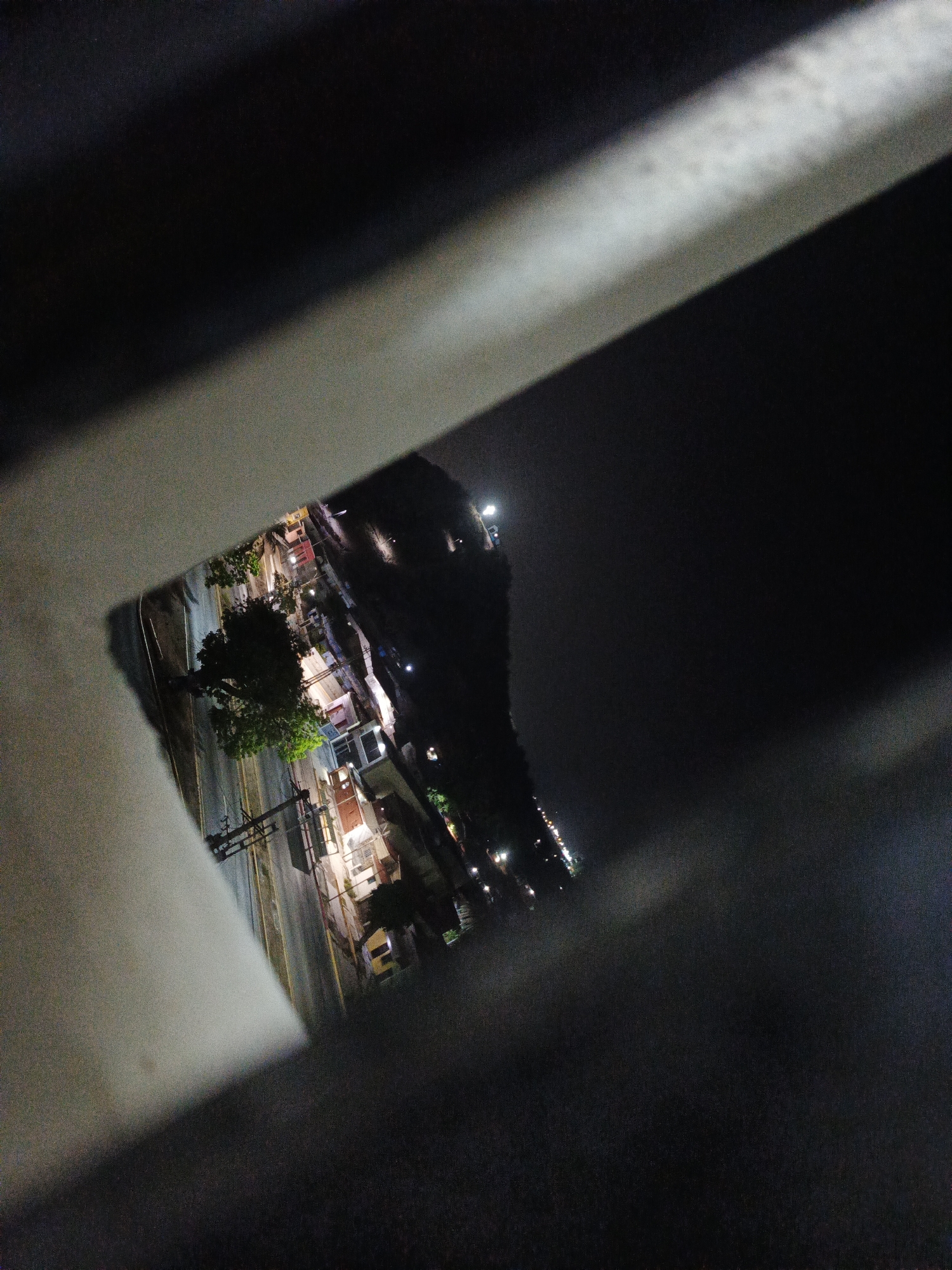 The image depicts a night scene in Los Robles, Venezuela, viewed through an obscured opening, likely between concrete or metal structures. The visible area contains a street with visible lane markings, overhead utility lines, and some vegetation, possibly a tree. There are buildings lining the street, illuminated by streetlights and interior lighting. Beyond this immediate street view, a dark and undefined background suggests a slope or hill.