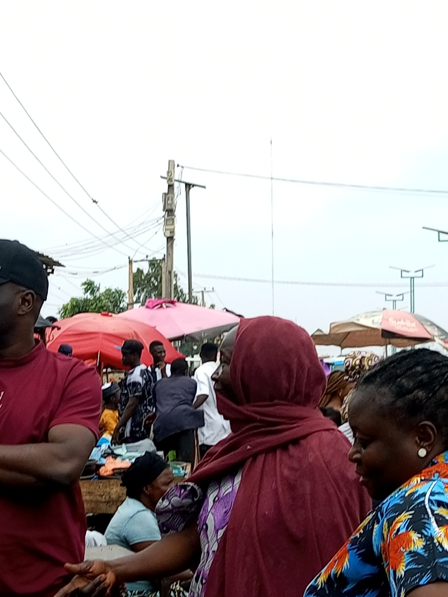 The image depicts a crowded outdoor market scene in Tempure, Nigeria. The foreground features the shoulders and back of a person wearing a maroon shirt and a black baseball cap, facing left. To the right, a woman with dark skin and braided hair, wearing a colorful patterned shirt and white earrings, is visible in profile, looking to the left. Behind them, several other individuals are seen, some obscured by umbrellas and stalls.
Dominating the background are several brightly colored umbrellas, including a red one and a pink one, suggesting various market stalls. Overhead, a network of electrical wires and poles crisscross the sky, indicating the presence of infrastructure within the urban environment. Solar-powered streetlights are also visible in the distance. The overall atmosphere appears to be that of a bustling marketplace, with people engaged in activities common to such a setting. The lighting suggests it might be daytime, possibly overcast.