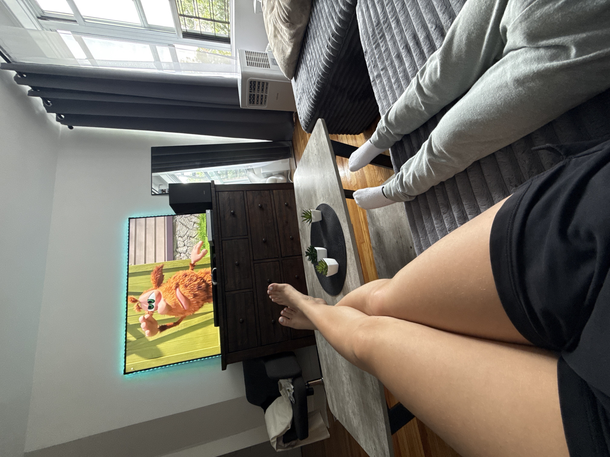 This is a first-person perspective, eye-level shot of two individuals, presumably a couple, relaxing in a living room. The image was captured at the 747 West 183rd Street, Apt 4A, New York, NY 10033, United States of America.
In the foreground, the legs of one person are crossed at the ankles, with bare feet visible. They are wearing black shorts. To the right, the legs of another person are extended, clad in light grey sweatpants and white socks. Both individuals are seated on a grey upholstered sectional sofa, with their feet resting on a textured grey rug.
A light grey coffee table sits between the sofa and a dark wood dresser. On the coffee table, there are two small potted succulents.
Behind the coffee table, a dark wood dresser holds a television set displaying an animated character. The TV is framed by green LED strip lighting. To the right of the dresser is a tall, slender black speaker. A full-length mirror stands behind the speaker.
Further to the right, a large window with white panes is visible, covered partially by dark grey curtains. Outside the window, greenery and trees are visible. An air conditioning unit sits on the floor below the window.
The room is furnished with a dark upholstered armchair with a cream-colored cushion to the left of the dresser. The walls are painted a neutral off-white. The floor appears to be hardwood.
The overall impression is a casual, relaxed domestic scene.
