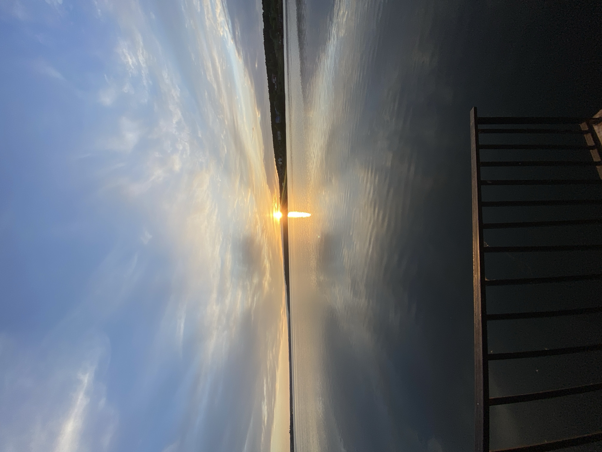 The image depicts a sunset over a body of water, possibly a lake or river. The sun is positioned on the horizon, reflecting brightly on the water's surface. The sky is a mix of blue and gray clouds, indicating either early morning or late evening. A strip of land is visible in the distance, suggesting the opposite shore. A dark, wooden railing or fence is positioned on the right side of the frame, likely part of a dock or viewing platform. Based on the given information, this scene could be from Votkinsk, Russia, located on the Votkinsk Reservoir. There is no evidence of people in the image.