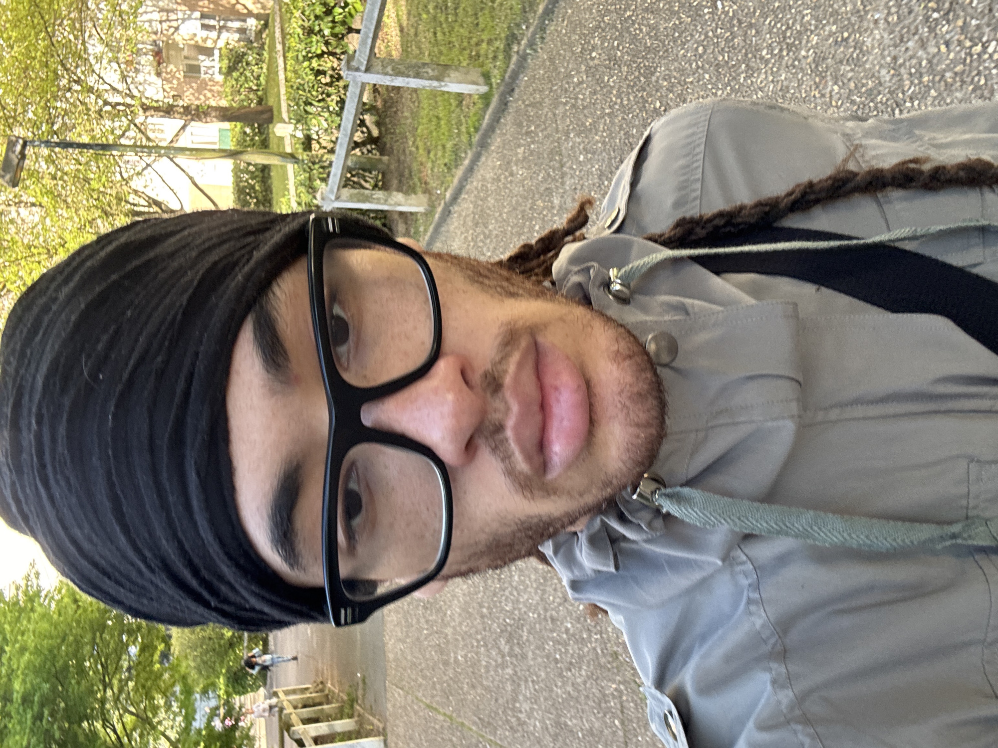 A close-up, slightly rotated, outdoor selfie depicts a person with brown skin, wearing a black turban, square black glasses, and a gray jacket. They have a short brown beard and dreadlocks. A black strap crosses their shoulder. In the blurred background, a sidewalk, grass, a fence, trees, and buildings are visible, suggesting an urban or suburban park setting. Given the prompt, the location is Les Ulis, France. A second person is walking away in the background.