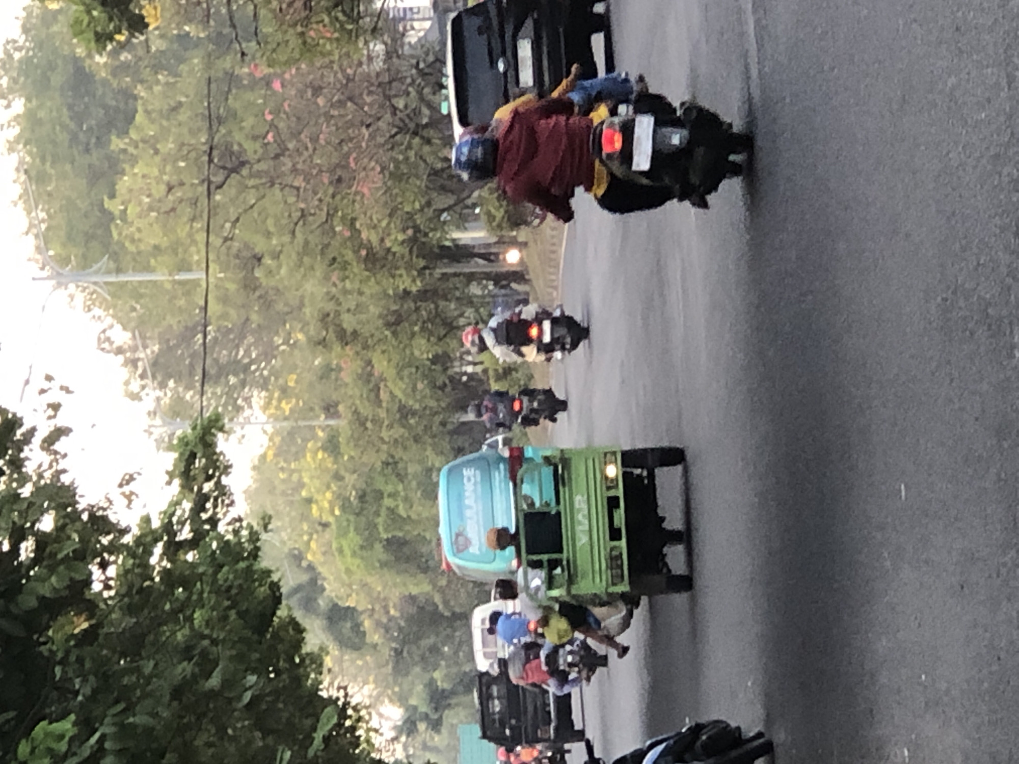 The image depicts a street scene in Sumurame, Indonesia, featuring a variety of vehicles and individuals navigating a road bordered by trees. In the foreground, a green, three-wheeled vehicle with the brand "VIAR" visible on its rear is prominent, carrying at least two passengers. To the left, a black pickup truck is visible. Further down the road, a light blue ambulance-like van with the word "AMBULANCE" written on its side is in motion. Several motorcycles are also present, with riders wearing helmets, including one in a red jacket and another in a darker jacket. In the background, more trees and a lamppost are visible, suggesting an urban or suburban setting. The lighting indicates it is daytime, possibly late afternoon due to the soft light and long shadows. The road surface appears to be asphalt.