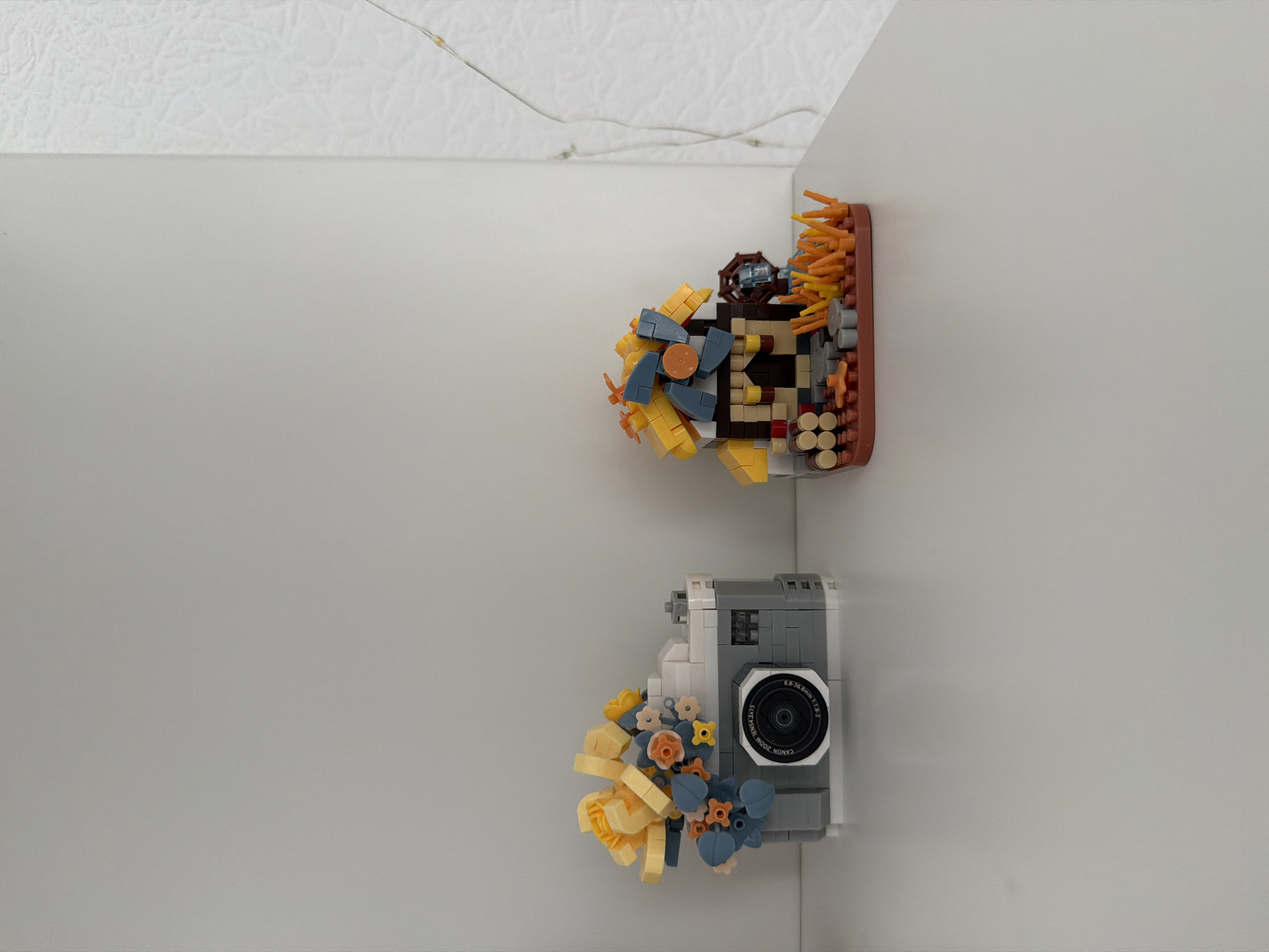 On a white, flat surface, two Lego constructions are displayed against a plain, light gray background. To the left is a Lego model of a camera, predominantly gray with white accents, decorated with yellow and blue floral elements. To the right is a Lego model of a small house with a thatched roof, featuring brown, yellow, and blue pieces, surrounded by logs and orange foliage. The background on the right transitions to a white, textured surface. The scene is well-lit, and there are no people or animals present. The location is not specified in the image or the provided context.
