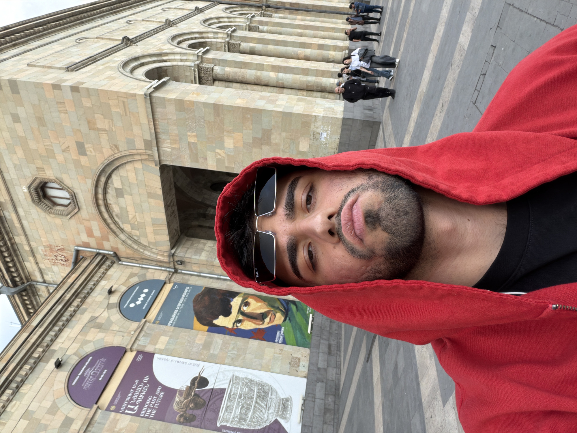 The image shows a man in a red hooded sweatshirt and sunglasses taking a selfie in front of the National Gallery of Armenia in Yerevan. Behind him are stone buildings with arches and columns, along with various posters displaying art and historical information. Some people are visible walking in the background. The paving stones on the ground show a striped pattern.