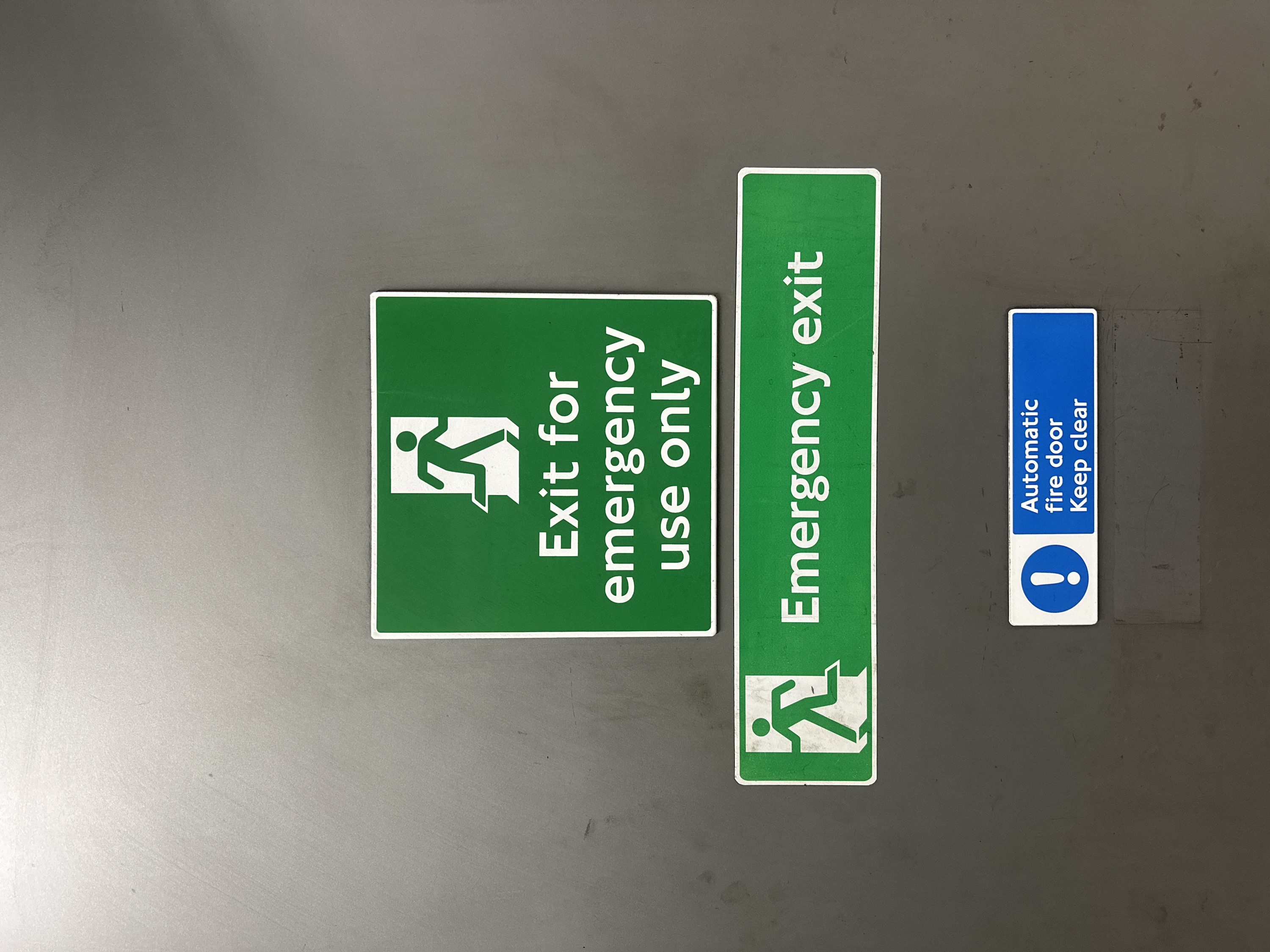 The image displays three horizontally aligned safety signs mounted vertically on a light grey, possibly metallic, wall or door, indicative of an interior location within the City of London, United Kingdom.
The top sign is rectangular with a green background and white border. It features a white pictogram of a running figure exiting a doorway above the white text "Exit for emergency use only."
Below it, the middle sign is also rectangular with a green background and white border. It shows a white pictogram of a running figure exiting a doorway to the left of the white text "Emergency exit."
The bottom sign is rectangular with a blue background and white border. On its left side, a white exclamation mark within a white circle is displayed. To its right, the white text "Automatic fire door Keep clear" is presented in two lines.
The grey surface exhibits minor surface imperfections, scratches, and dust. No individuals, actions, or specific interactions are depicted.