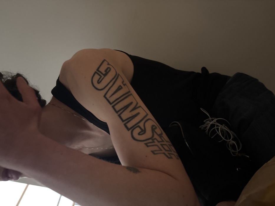 An individual with light skin and dark, curly hair is shown from a low angle. They are wearing a black top and have a tattoo on their arm that reads "SWAG." One hand is near their face, while the other arm is closer to the camera. A thin necklace is visible around their neck. A black jacket with a zipper and entangled white headphones are visible to the right of the image. The background is a dimly lit, neutral color.