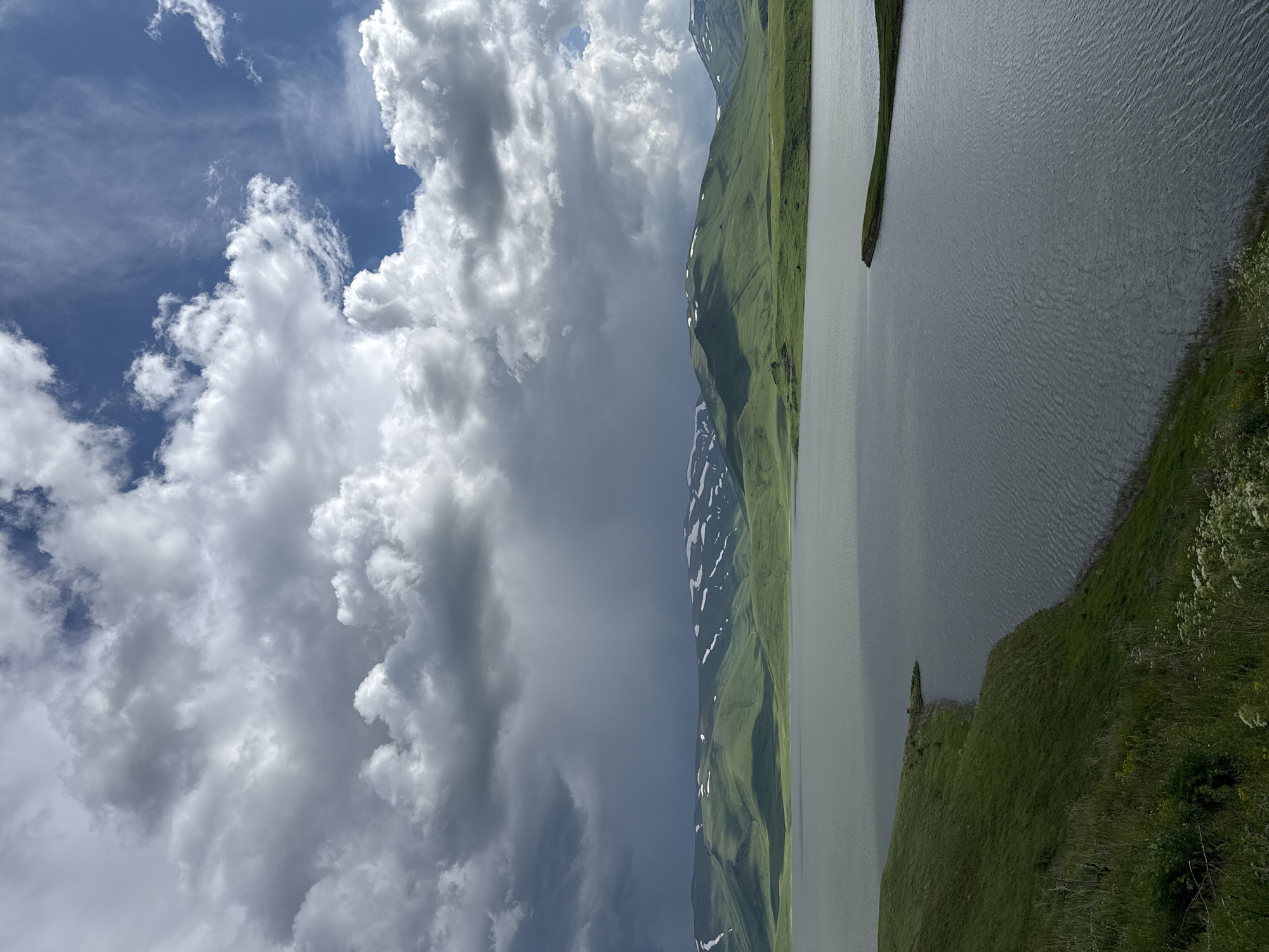 The image depicts a serene landscape featuring a tranquil lake in the foreground, with rolling green hills and snow-capped mountains in the background. The sky is a dramatic canvas of contrasting clouds, with patches of blue sky visible amidst heavy, dark storm clouds on the left and lighter, fluffy white clouds on the right. The lake's surface is rippled, reflecting the sky. The foreground shows a grassy embankment leading down to the water. The location is identified as Tsghuk, Armenia, suggesting a natural, mountainous terrain. No people or specific actions are visible, creating a sense of stillness and natural beauty.