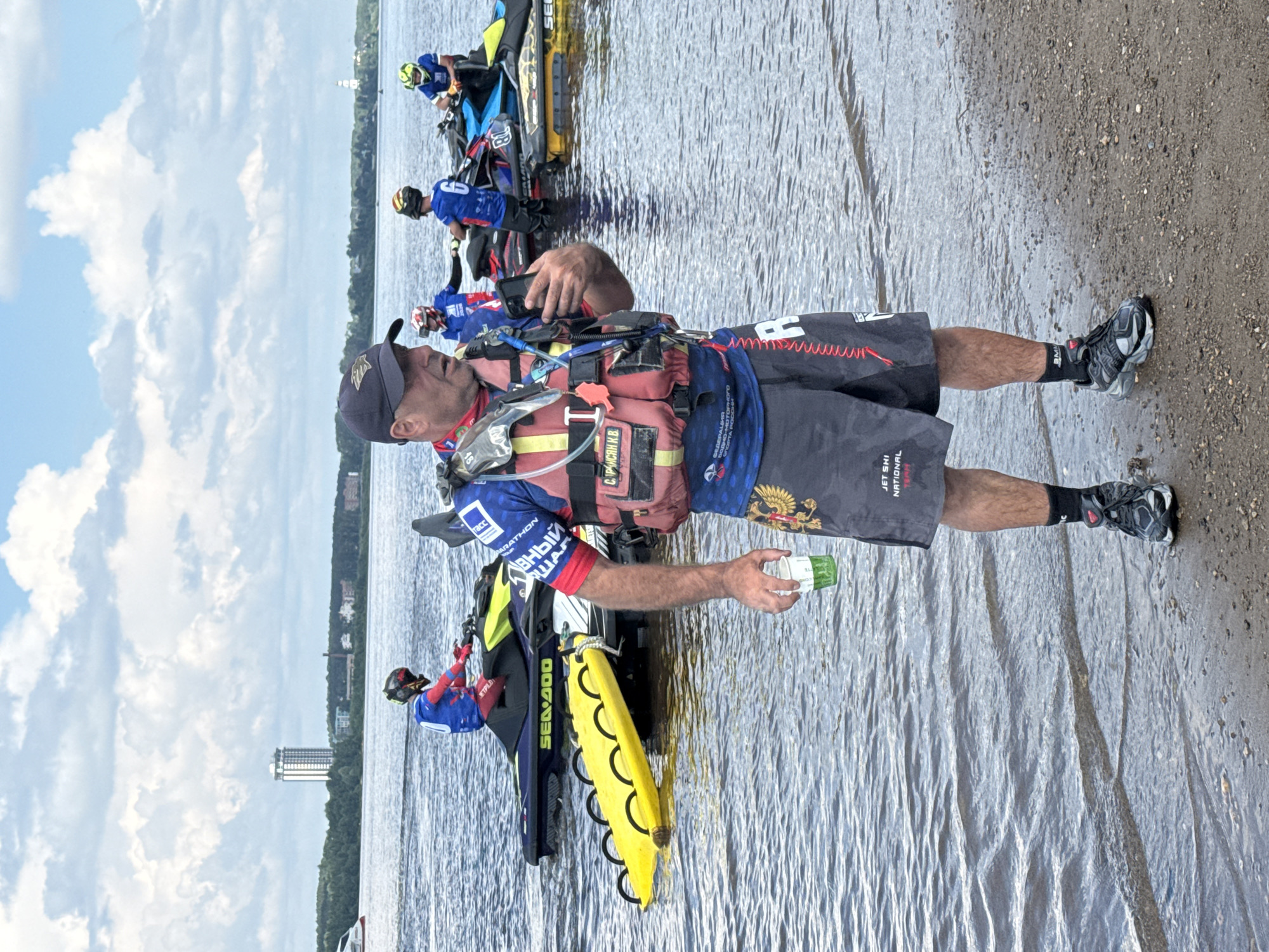 A man in a life vest and athletic shorts stands on a pebble beach, holding a drink and a cell phone. Behind him, several jet skis are parked in shallow water. In the background, a city skyline with a tall building is visible across a wide body of water. The man is wearing a dark baseball cap, a blue and red rash guard with text and logos, and gray shorts with a red and white design. He has on black athletic socks and gray and black sneakers. The jet skis are in various colors, including yellow, blue, and black. The water appears calm, and the sky is partly cloudy. This scene is likely the Kama River embankment in Perm, Russia, given the presence of the tall building which resembles the "Permskaya Vysota" residential complex.